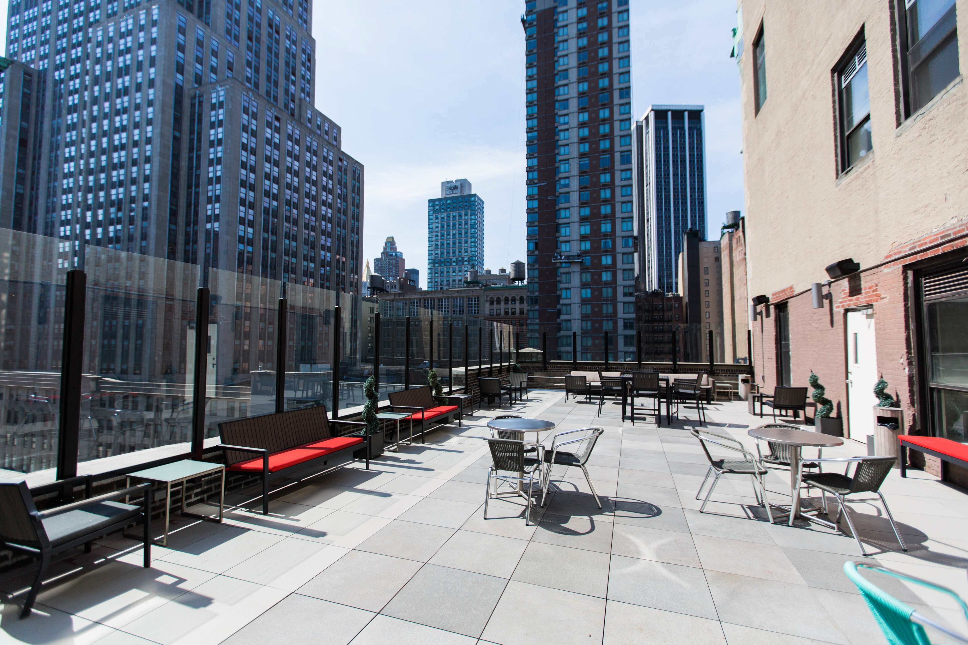 The image shows a rooftop terrace with seating areas and a view of tall buildings in an urban skyline.