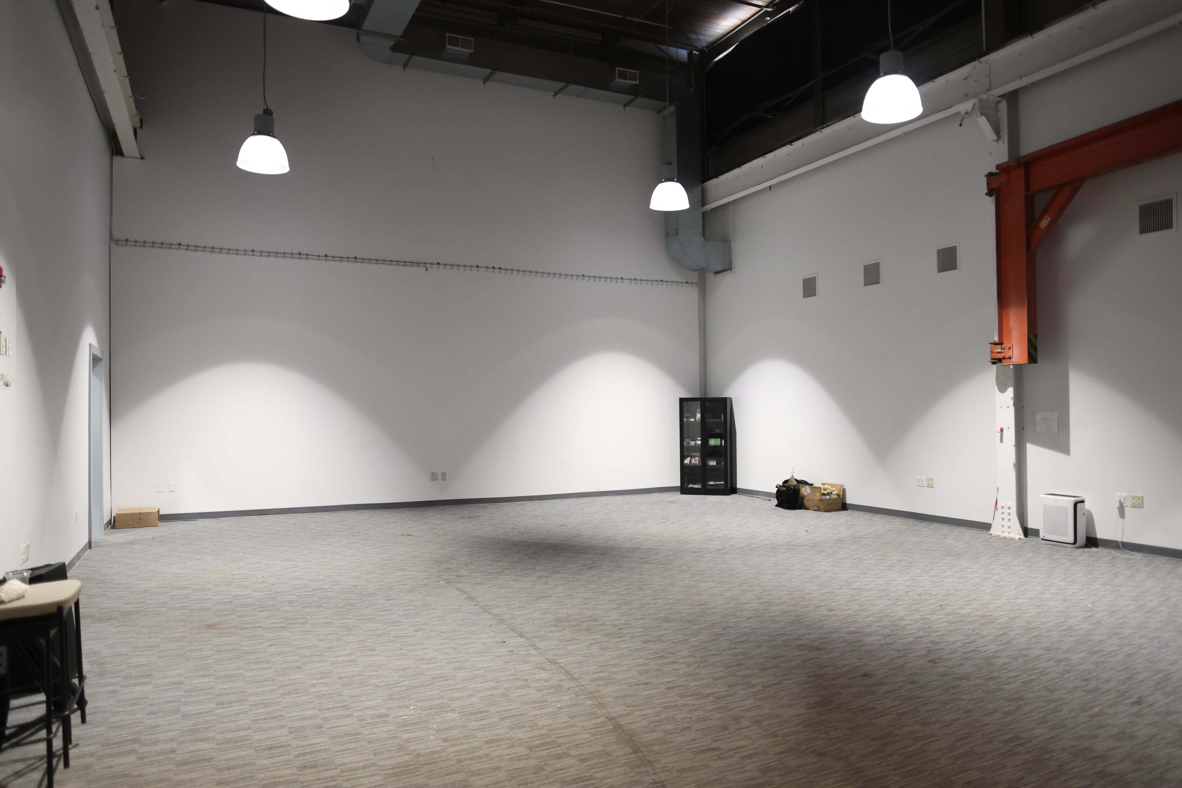 The image shows a large, empty room with gray carpeting, bare walls, and industrial-style lighting.