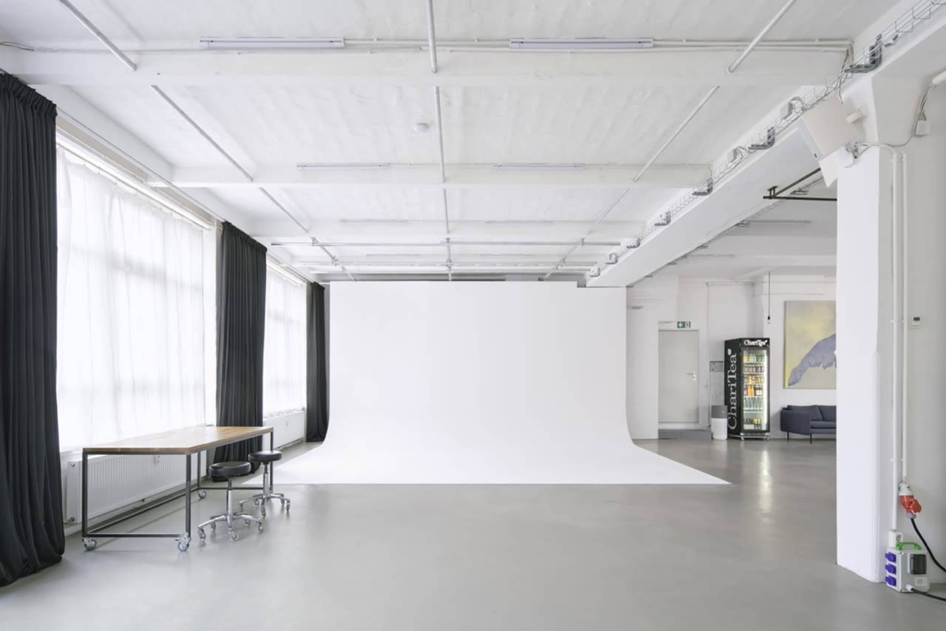 The image shows a spacious, well-lit studio with a large white backdrop, a wooden table with a chair on one side, and a vending machine against the wall.