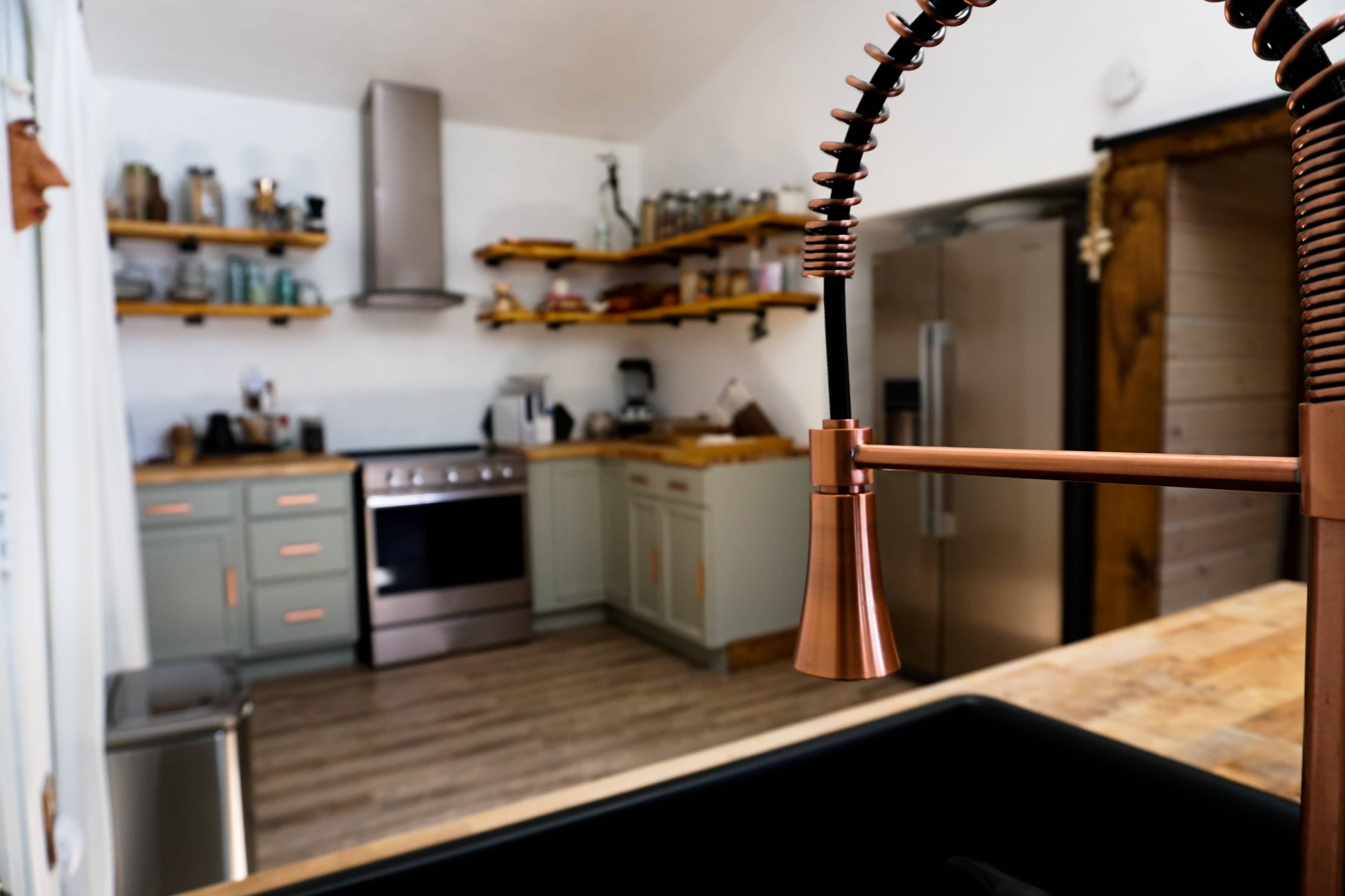 A modern kitchen with green cabinets, wooden shelves, and stainless steel appliances, featuring a prominent copper faucet in the foreground.