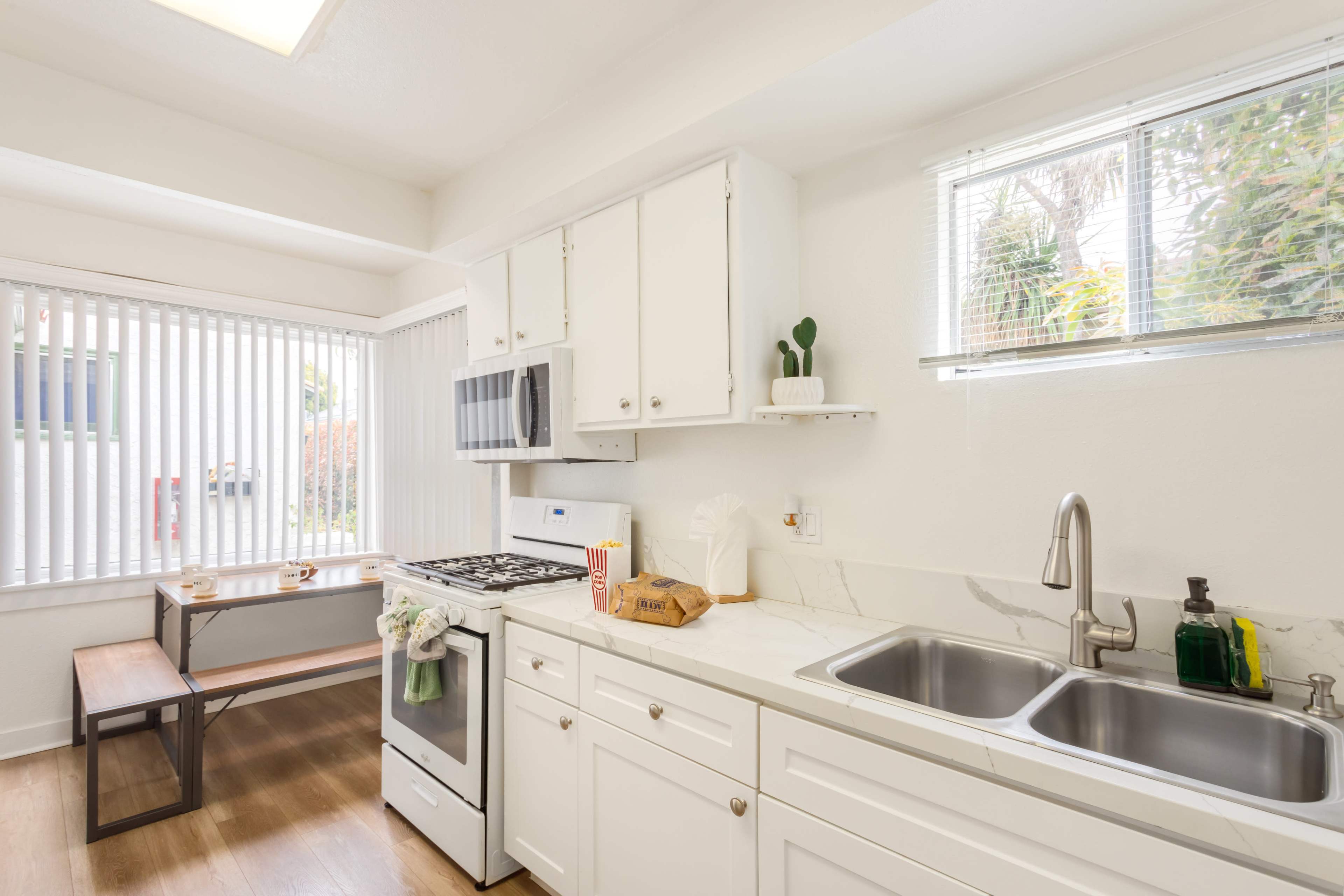 The image shows a modern kitchen with white cabinets, a stove, a sink, and a small dining table by a window with vertical blinds.