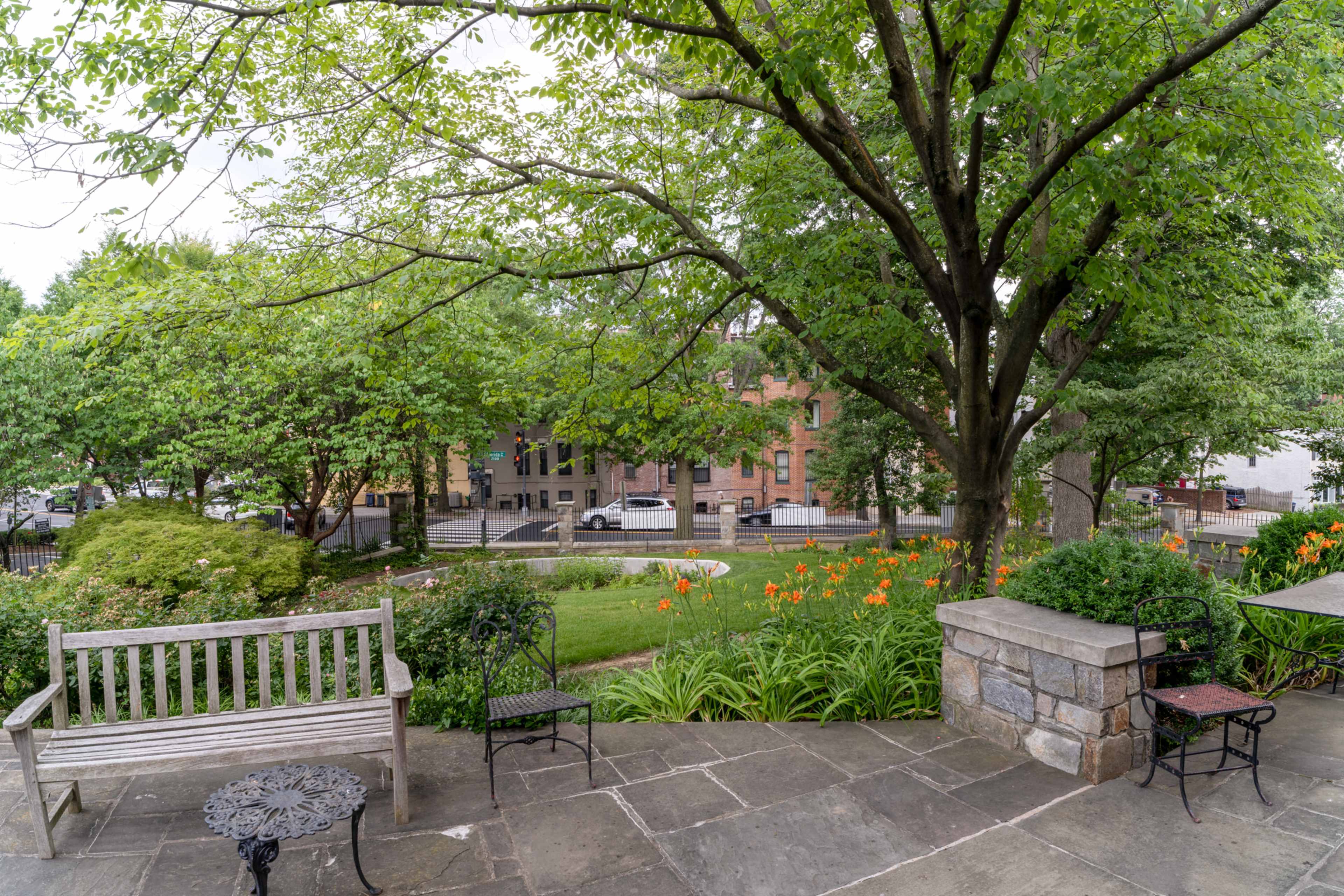A stone patio with a wooden bench and a wrought-iron table overlooks a grassy area adorned with orange flowers and large trees, with a street and buildings visible in the background.