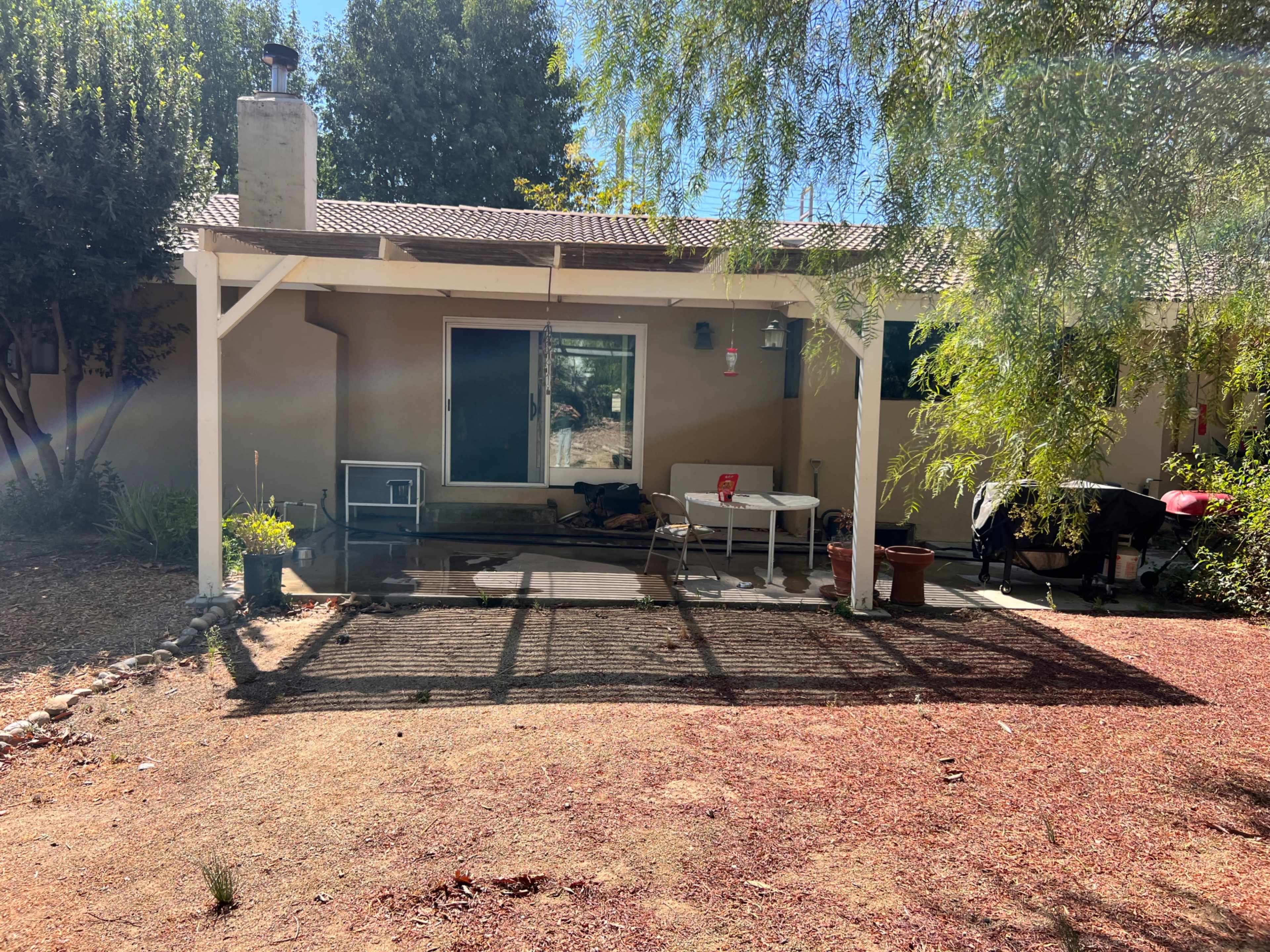 The image shows a patio area of a house with a covered seating space, a few potted plants, and a grill.