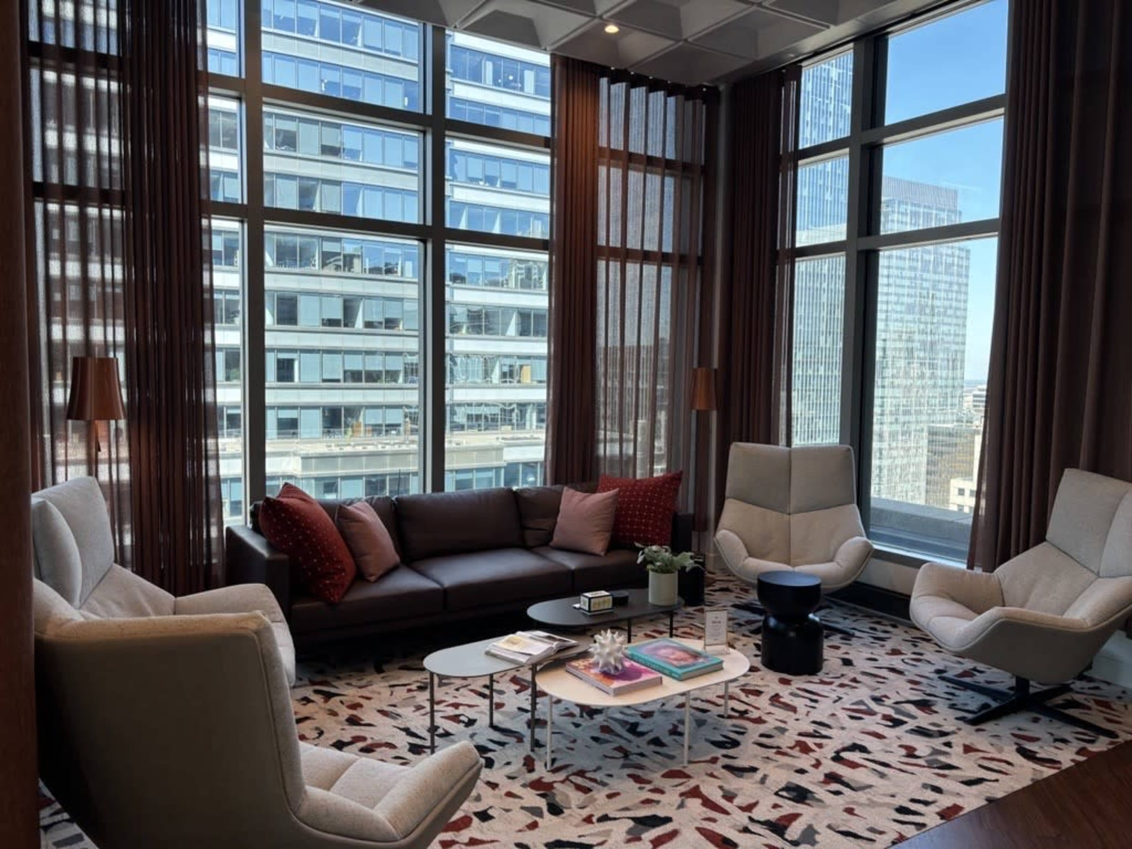 A modern lounge area features a brown sofa, two light-colored armchairs, a coffee table, and large windows displaying a cityscape.