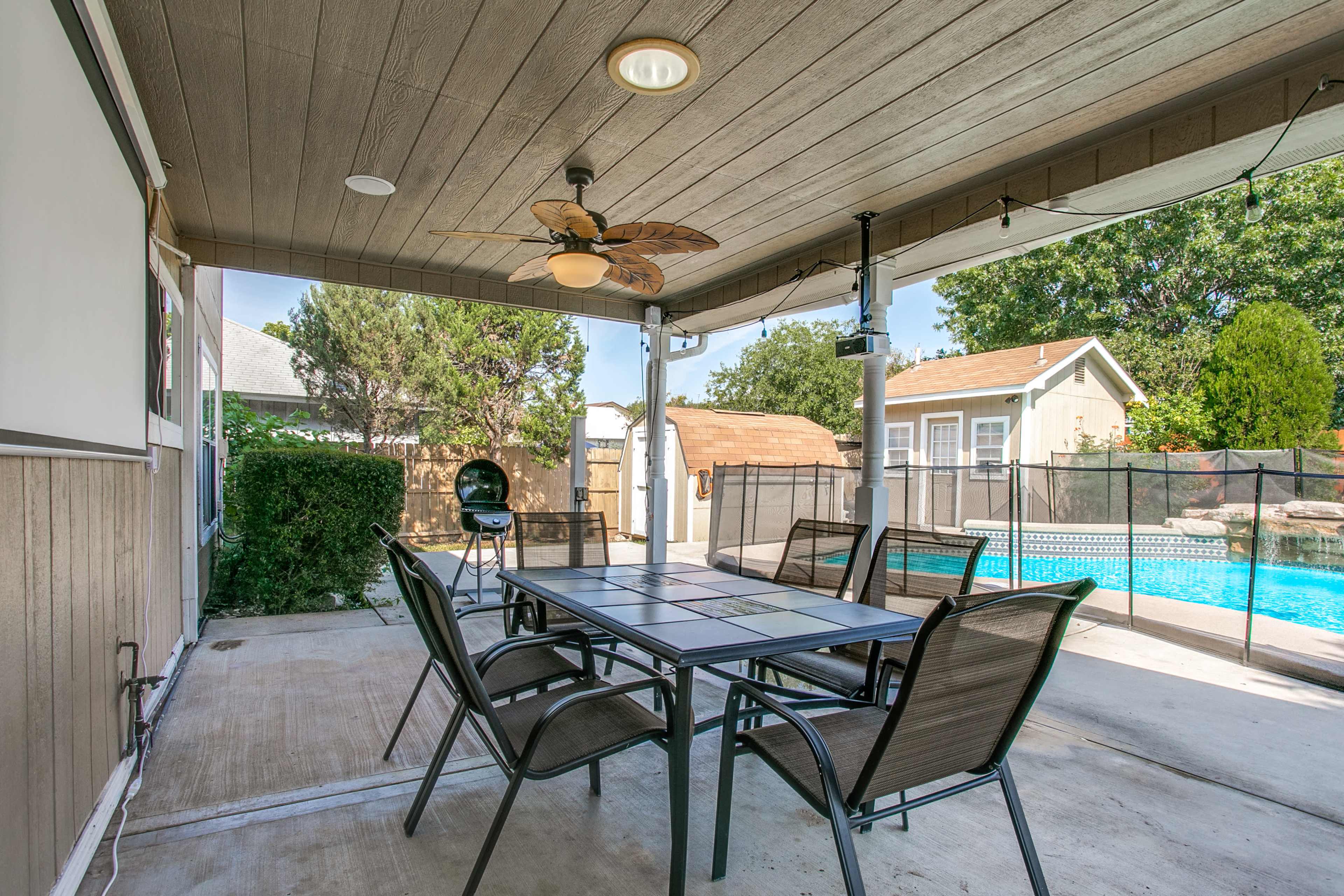 A covered patio area with a dining table and chairs, a ceiling fan, and a view of a swimming pool surrounded by a safety fence.