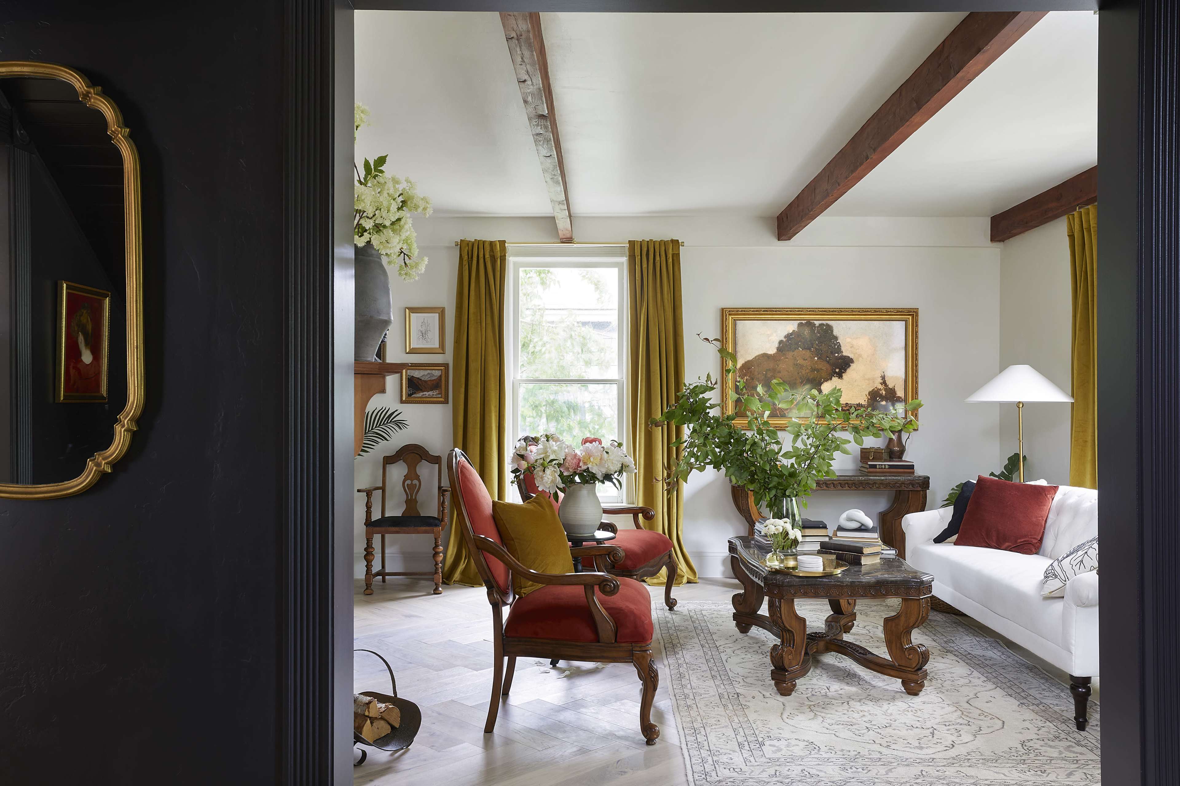 The image shows a living room with a white sofa, red accent chairs, a dark wooden coffee table, and large windows dressed with yellow curtains, surrounded by decorative plants and artwork.