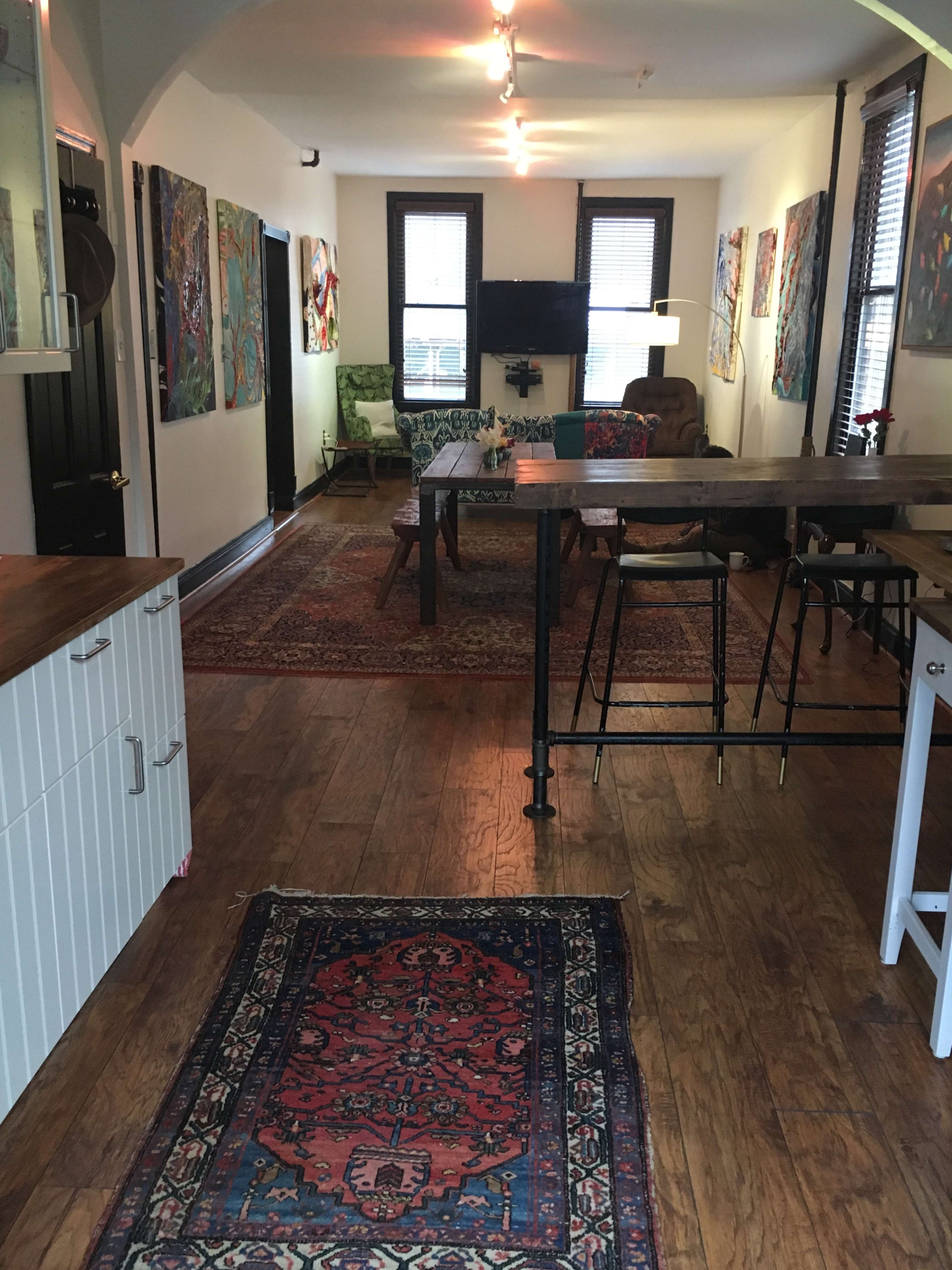 A colorful living space featuring a wooden dining table, several pieces of artwork on the walls, and a patterned area rug.