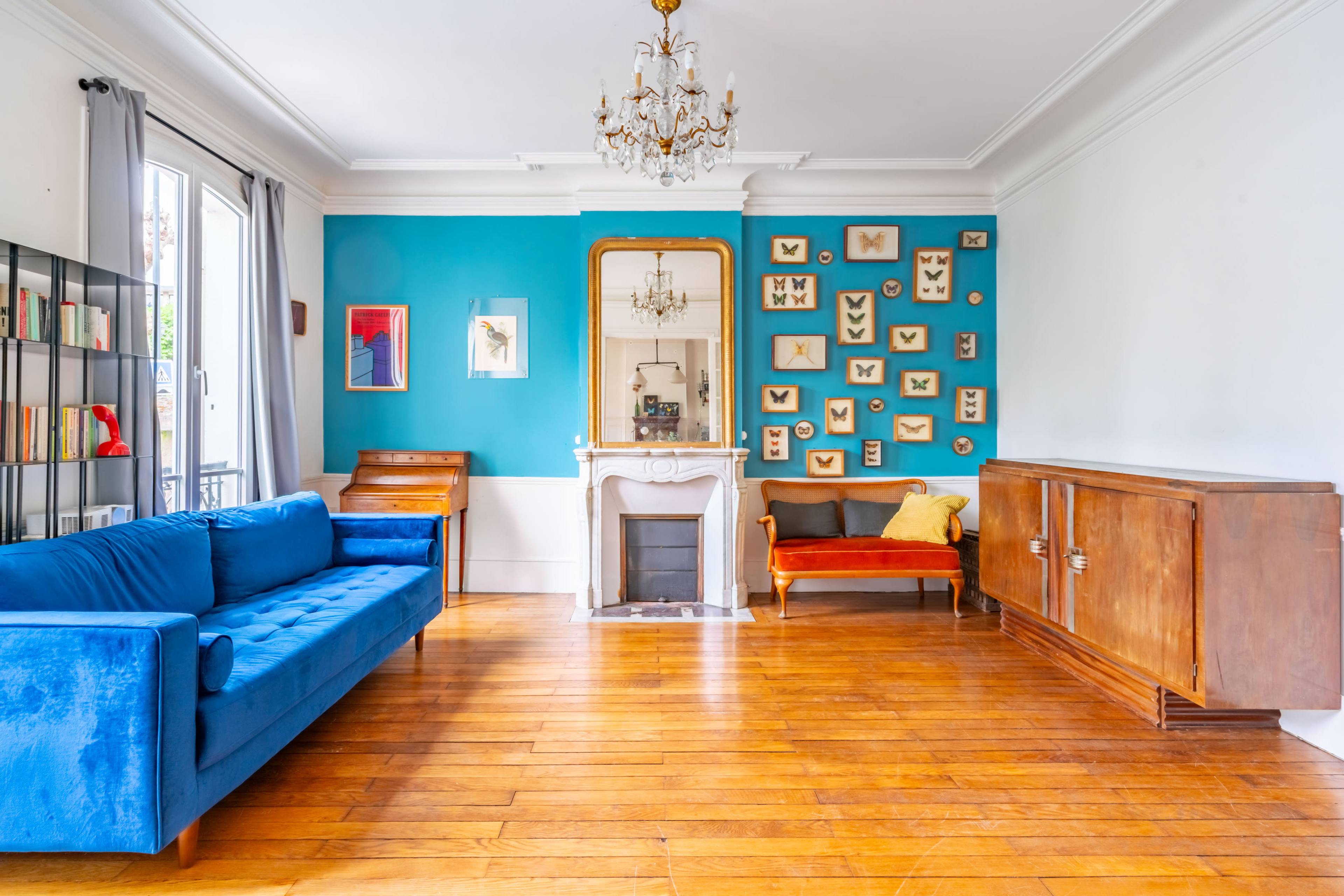 The living room features a blue sofa, a vintage armchair, a white fireplace, and a wall adorned with framed butterfly displays against a teal backdrop.