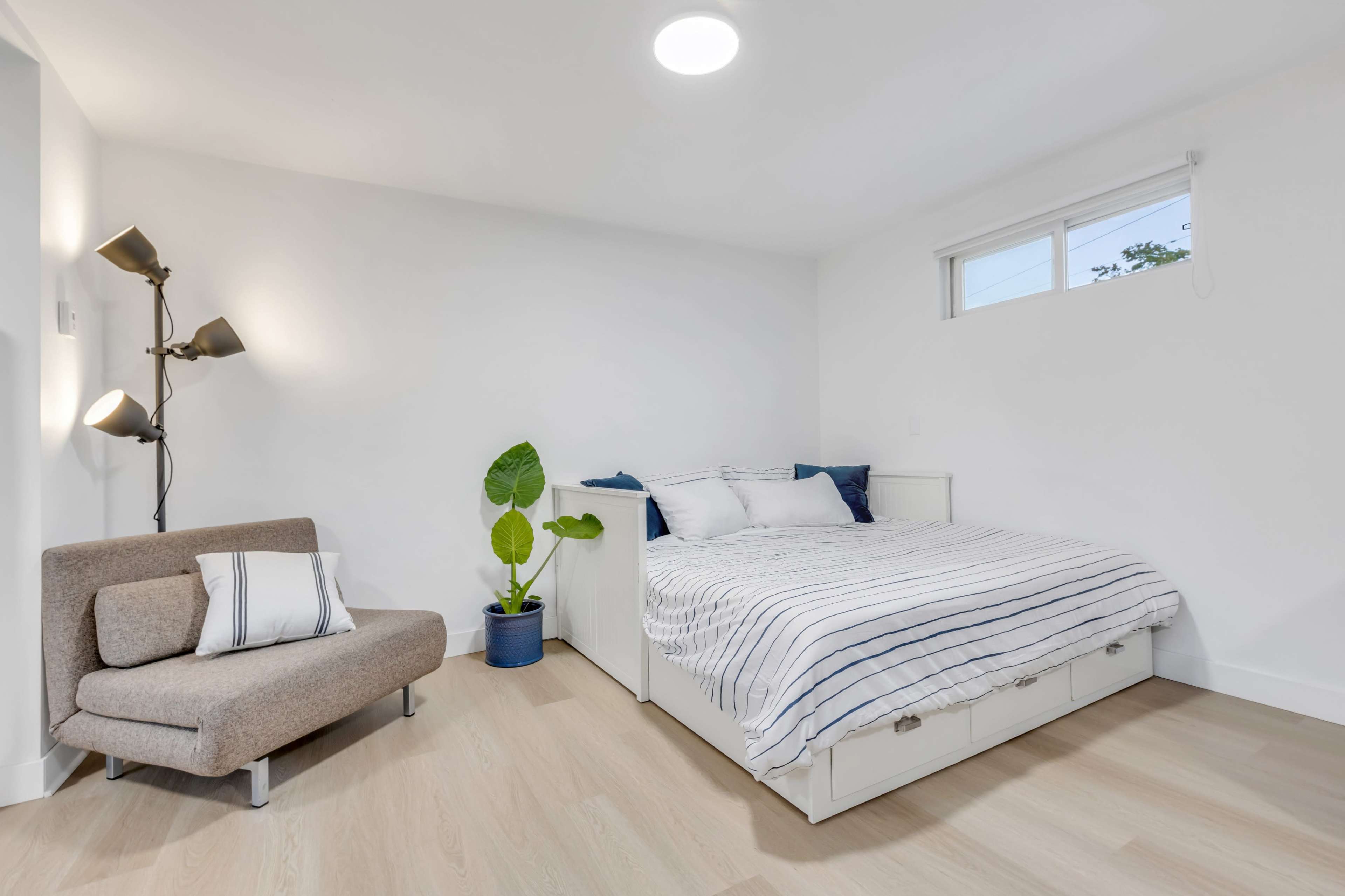 A modern bedroom featuring a bed with striped bedding, a small seating area with a gray chair, and a green plant in a blue pot.