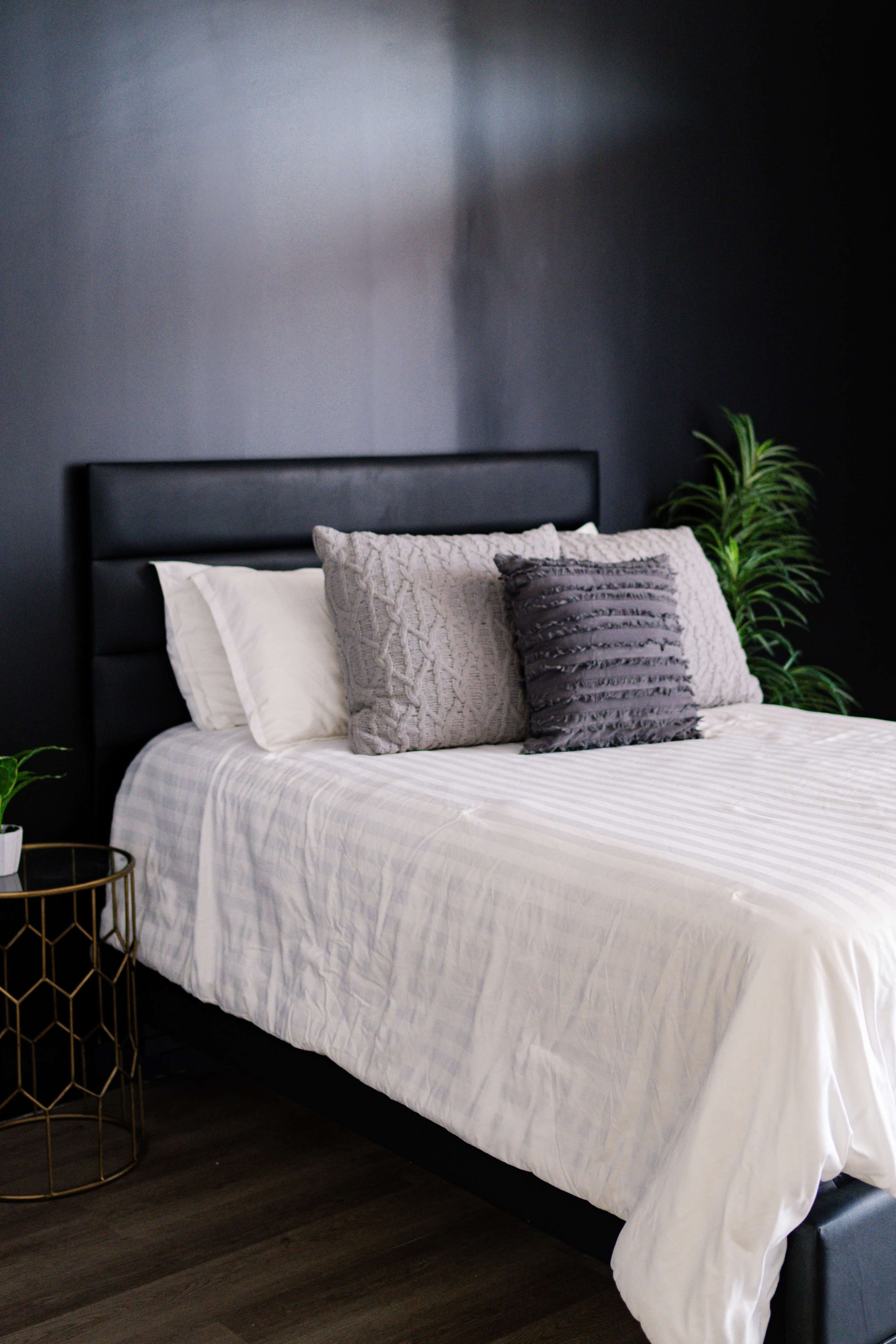 A neatly made bed with a black headboard is positioned against a dark wall, accompanied by a small gold side table and a potted plant.