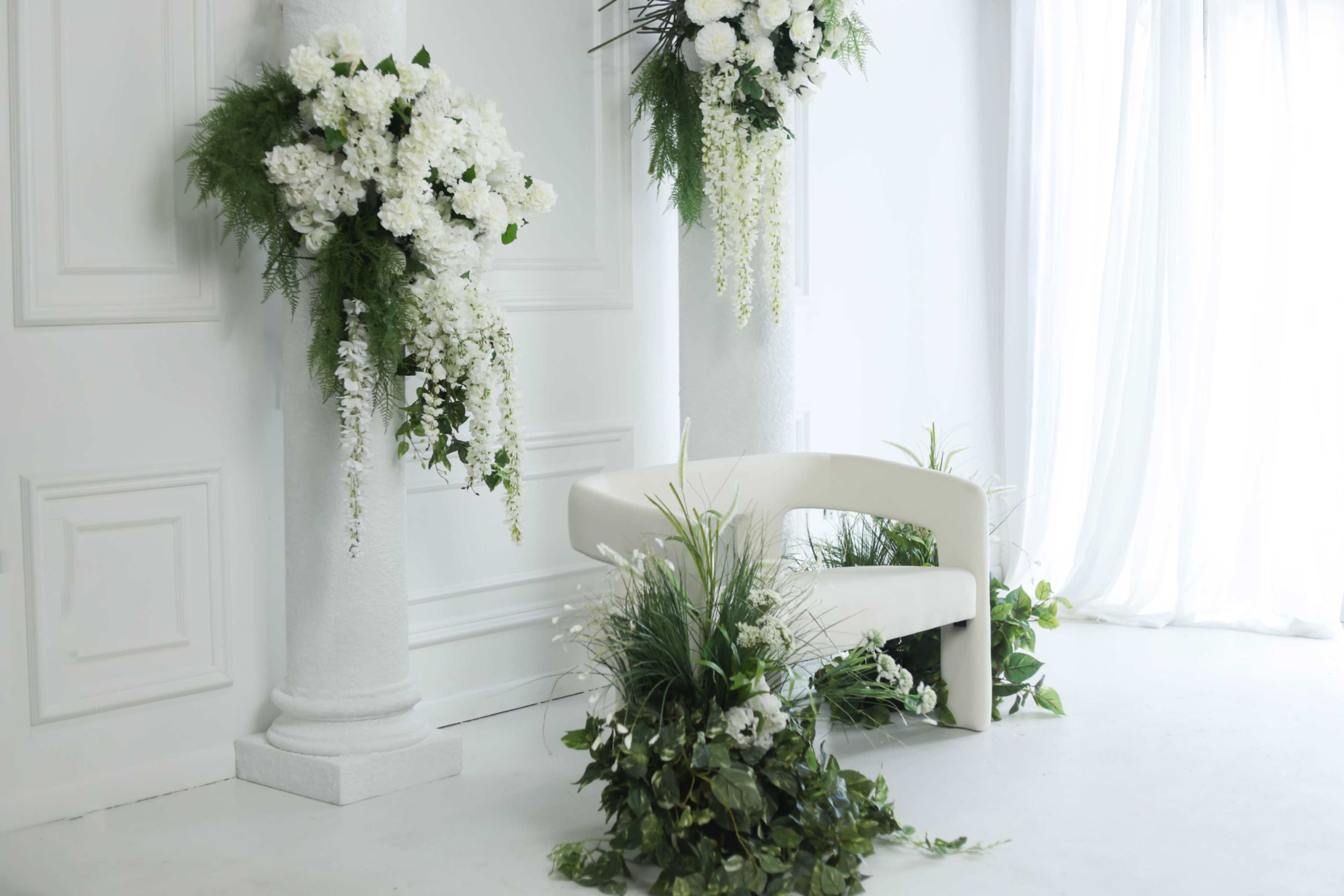 A minimalist white-walled space features a modern curved bench surrounded by large floral arrangements and greenery.
