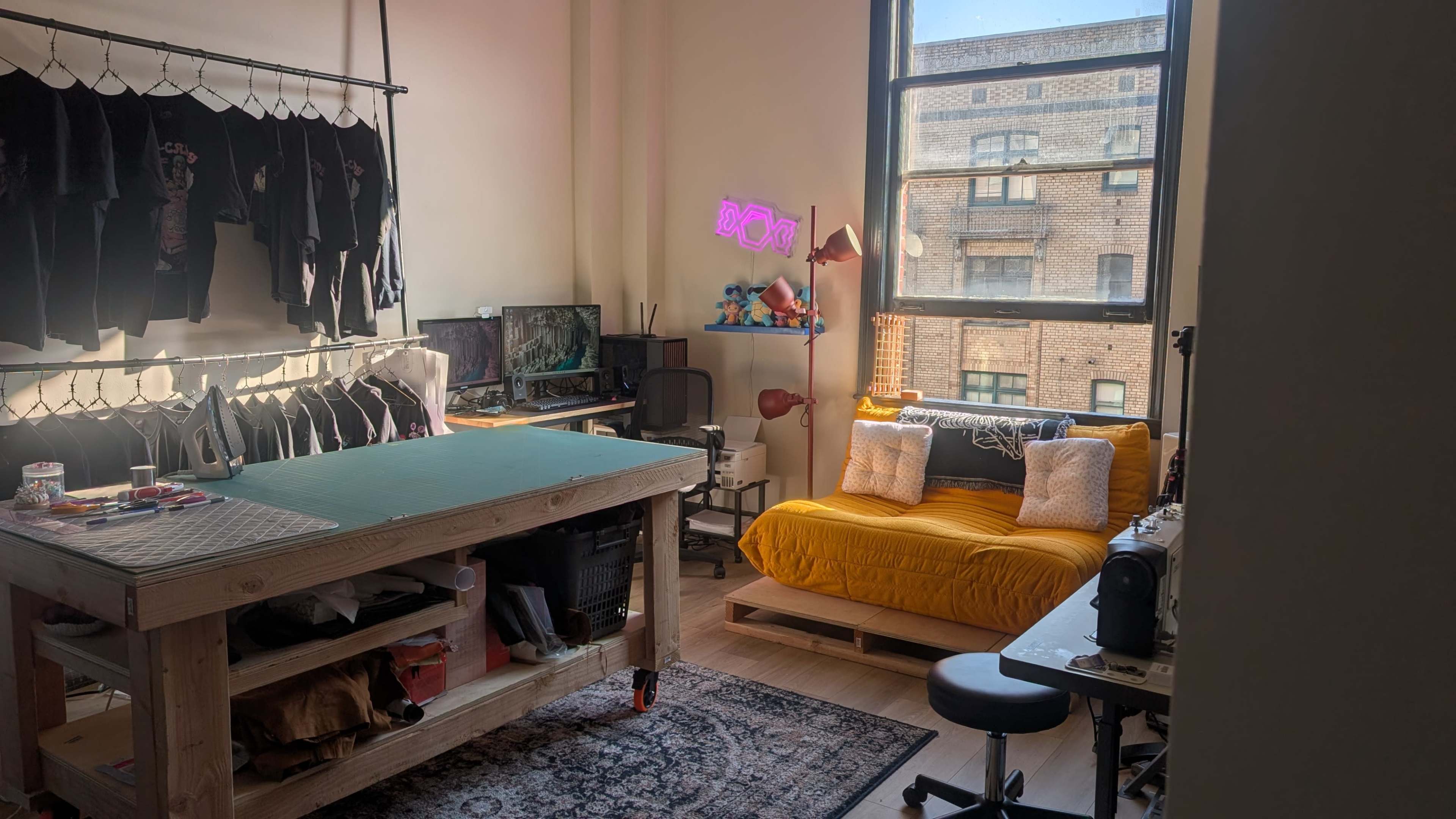 The image shows a brightly lit studio space featuring a large cutting table, a yellow couch with decorative pillows, and a view of a building outside the window.