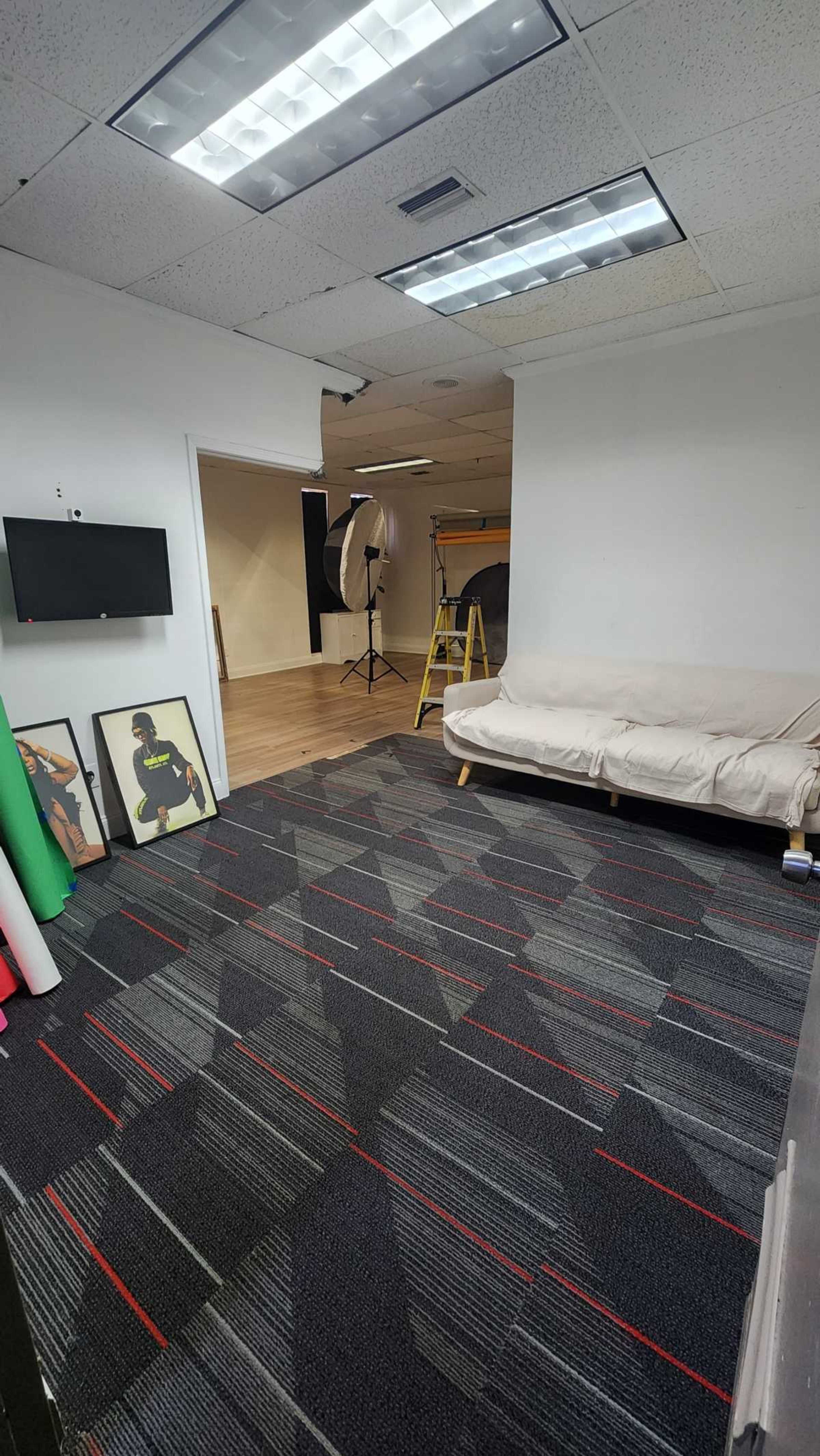 The image shows a spacious studio room with a patterned carpet, a white couch, a wall-mounted TV, and photography equipment set up in the background.