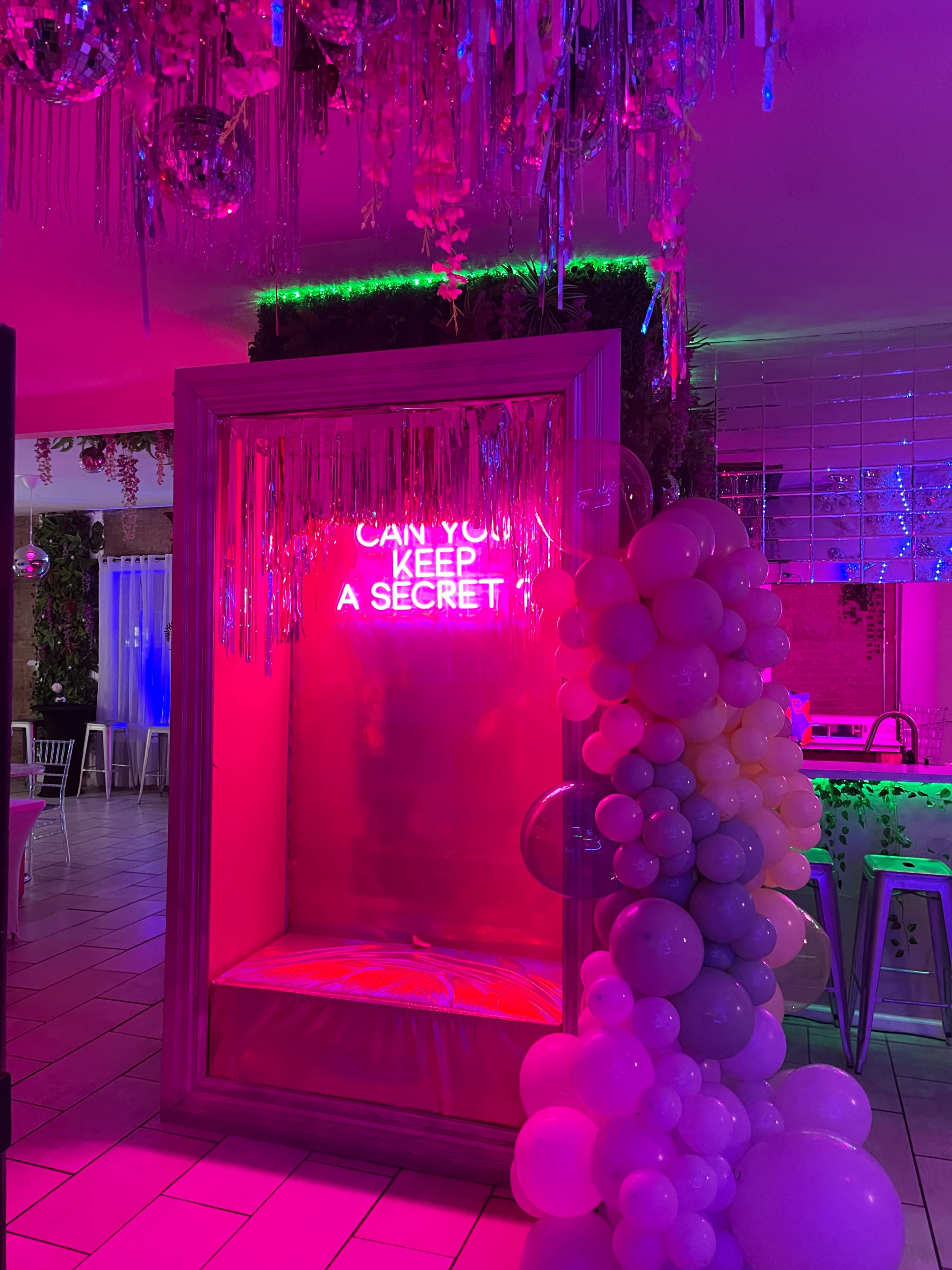 The image shows a brightly lit, decorated space featuring a neon sign that reads "CAN YOU KEEP A SECRET" inside a large frame, with colorful balloons and hanging decorations around it.