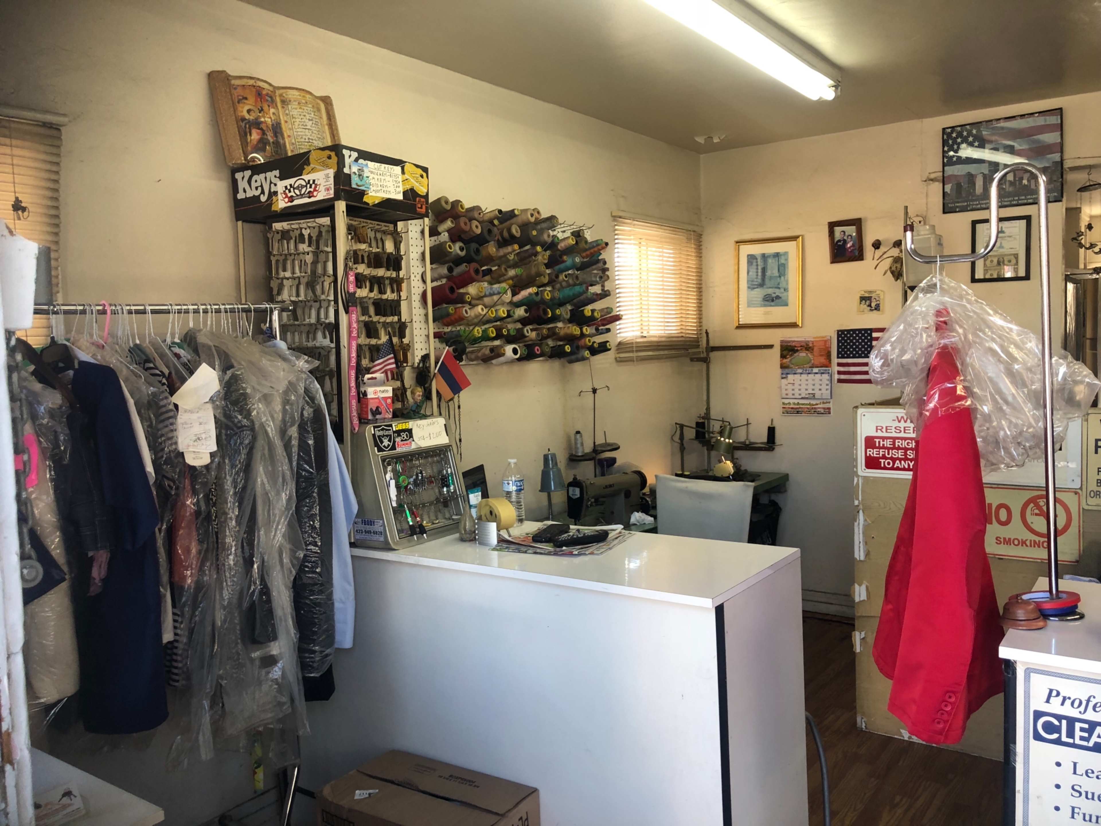 The interior of a small tailoring shop with a front counter, a rack of cleaned garments, and a wall adorned with spools of thread and various supplies.