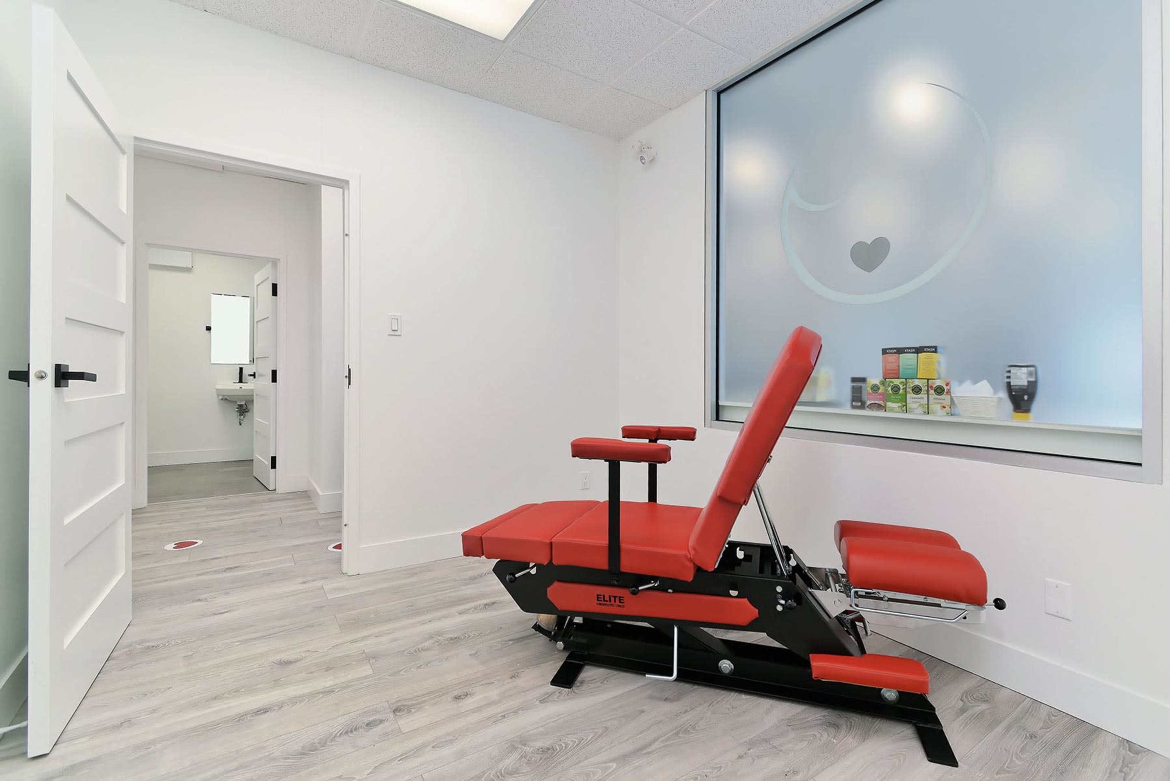 A bright, minimalist treatment room featuring a red therapy chair next to a large window, with a doorway leading to a bathroom visible in the background.