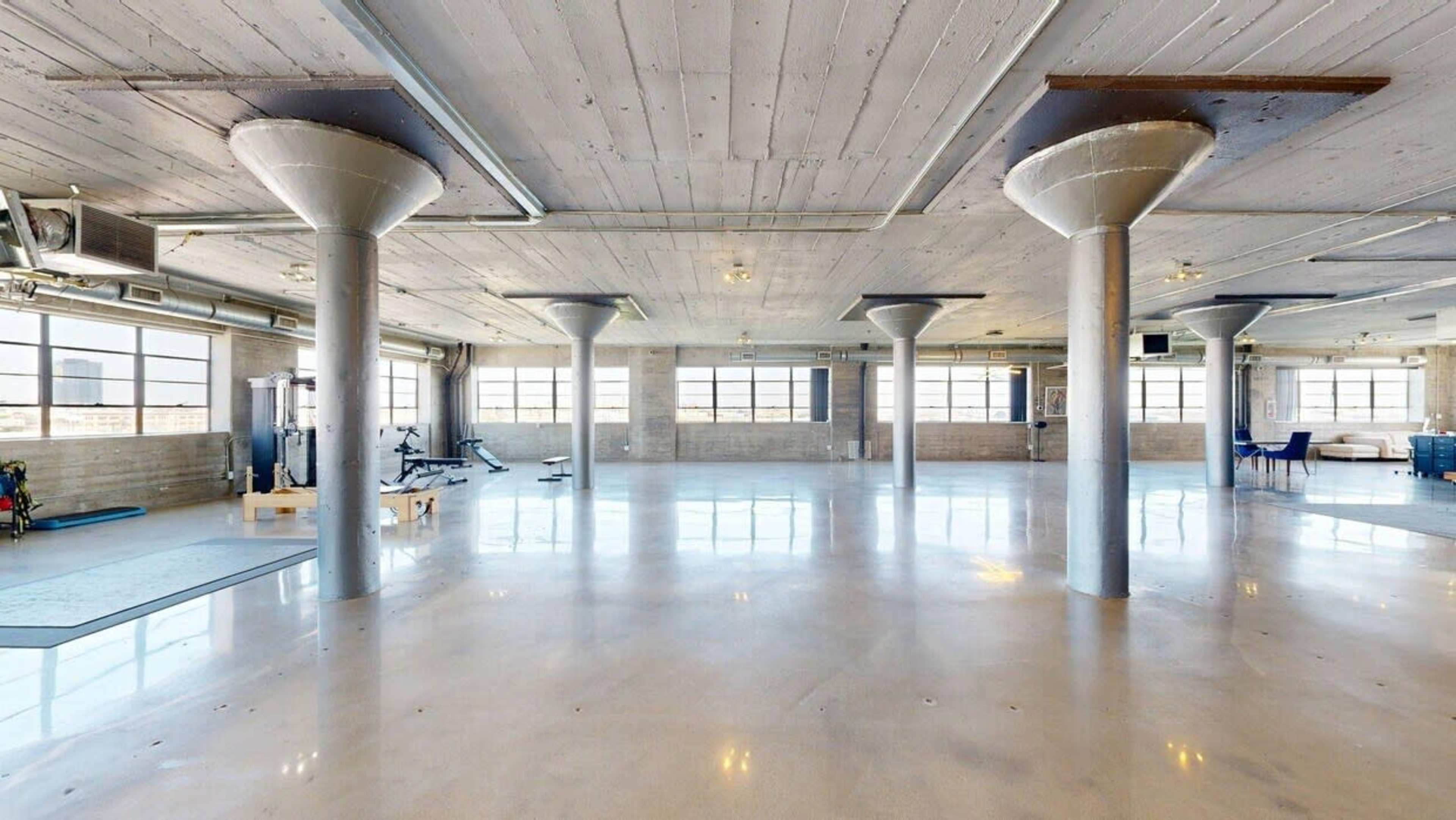 The image shows a spacious, modern gym area with polished concrete floors and large windows, featuring exercise equipment and pillars supporting the ceiling.