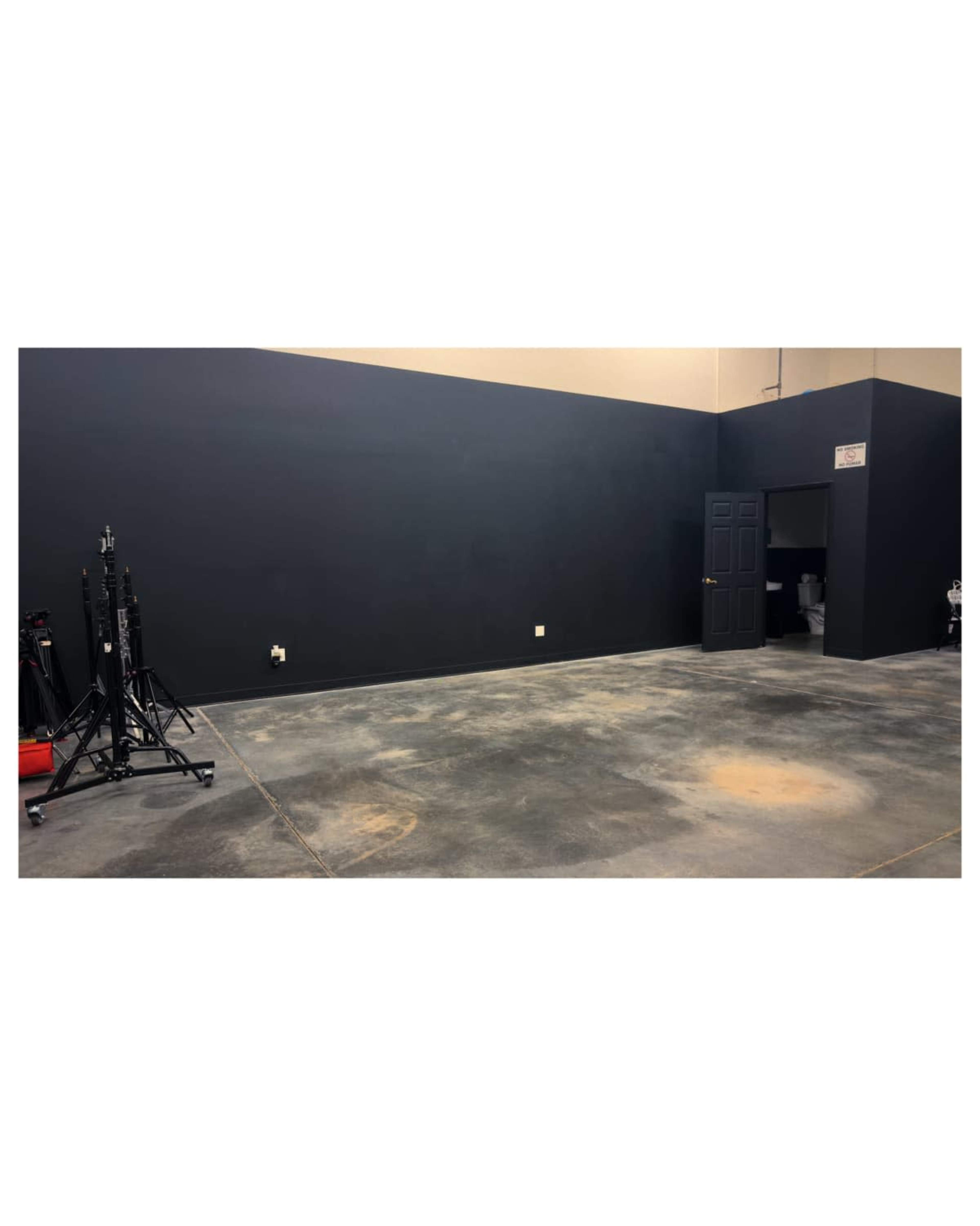 The image shows a large, empty room with black walls, concrete flooring, and a closed door at the far end.