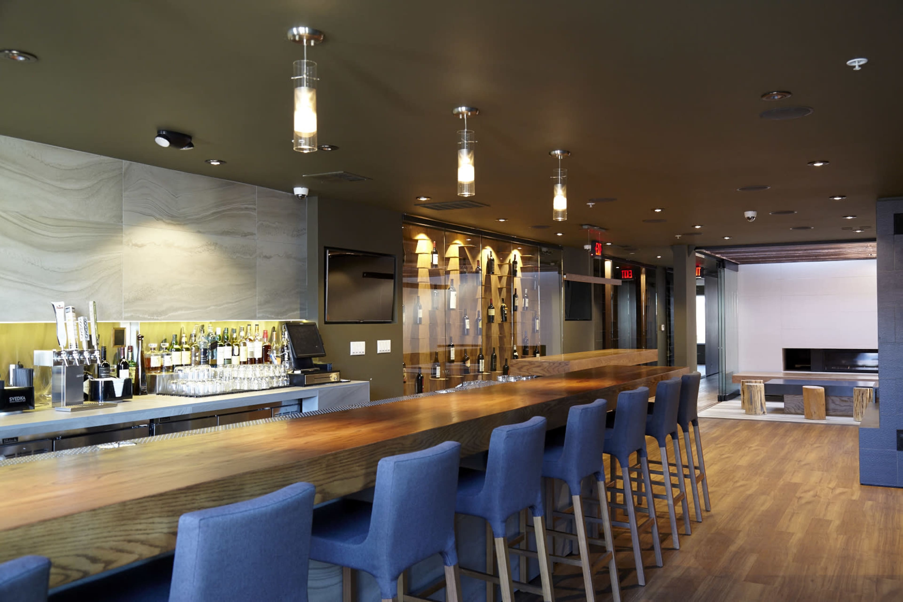 A modern bar area features a long wooden counter with high stools, shelves of drinks, and soft lighting.