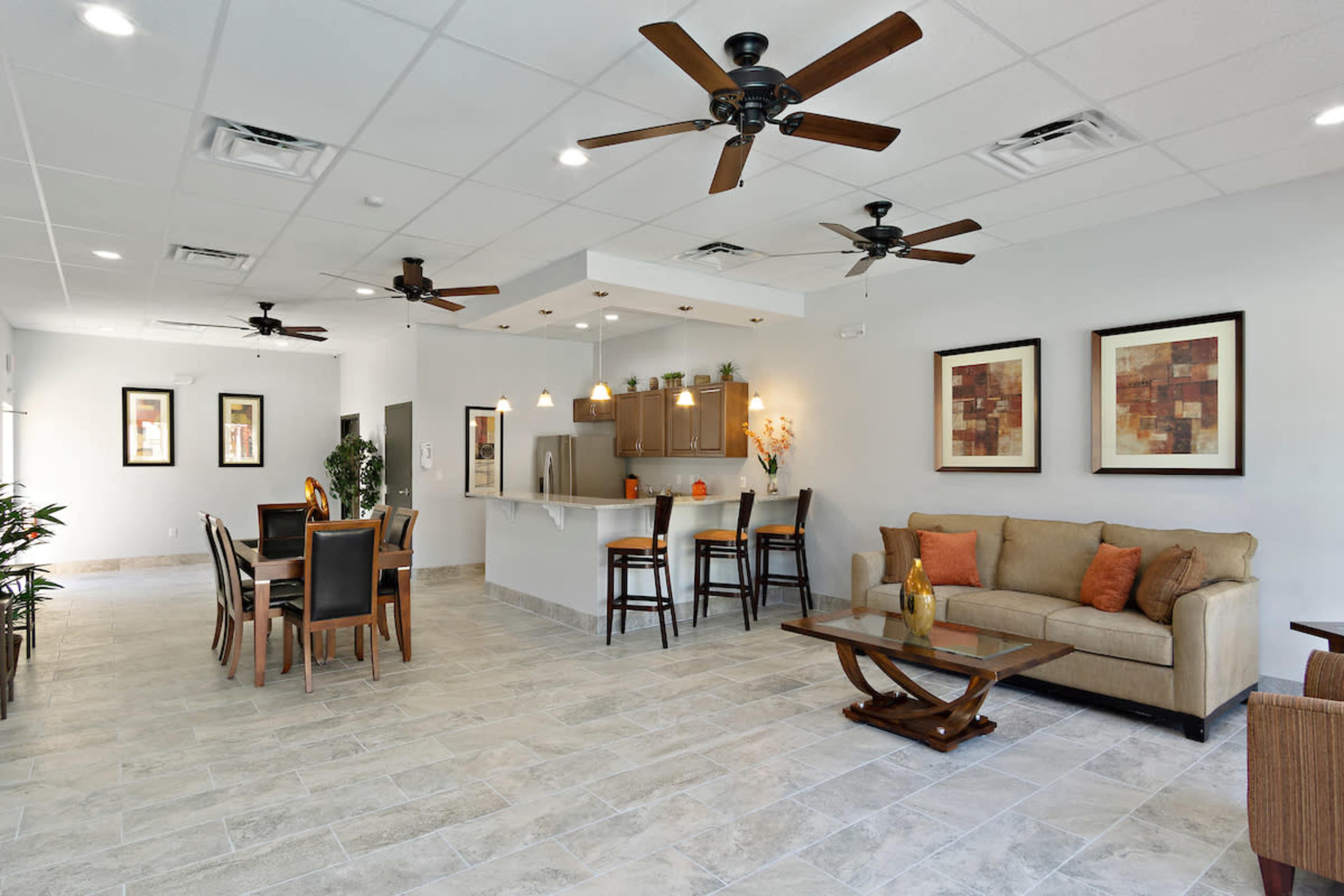 A modern common area with a beige sectional sofa, a dining table, and a kitchen bar, all set in a brightly lit space with ceiling fans and decorative artwork on the walls.