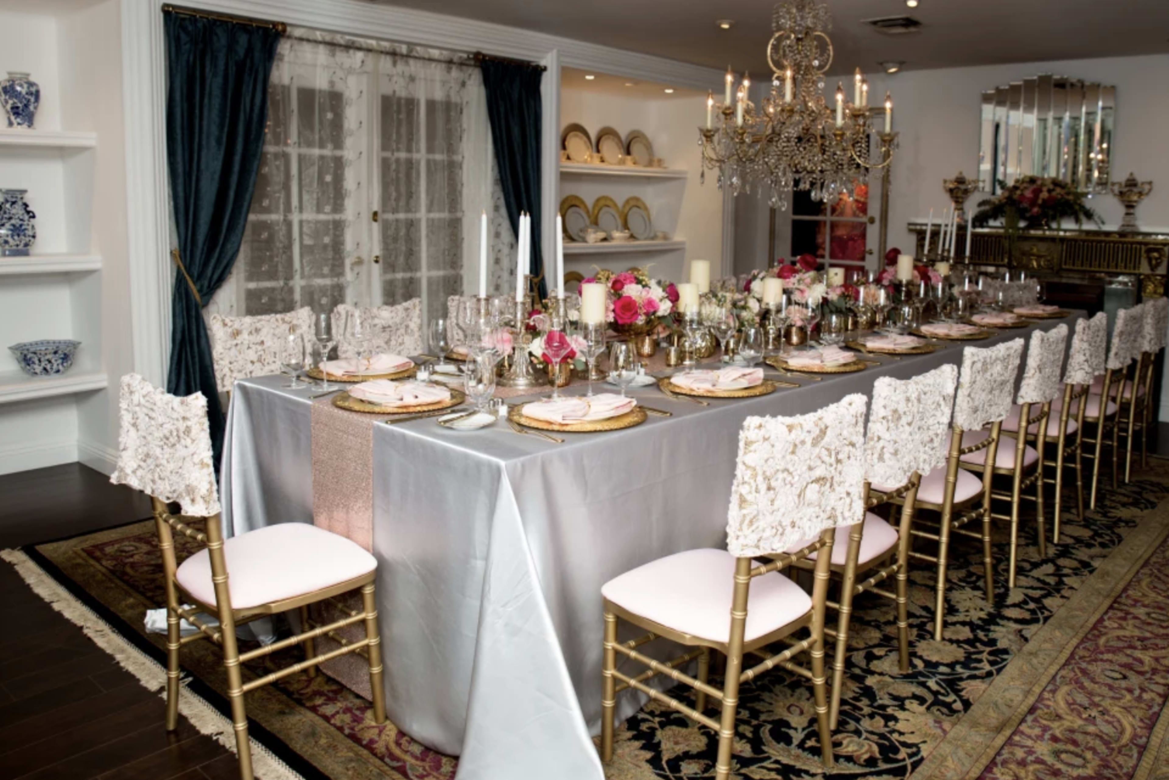 A long, elegantly set dining table with ornate chairs and floral centerpieces is arranged in a warmly lit room adorned with decorative plates and a chandelier.