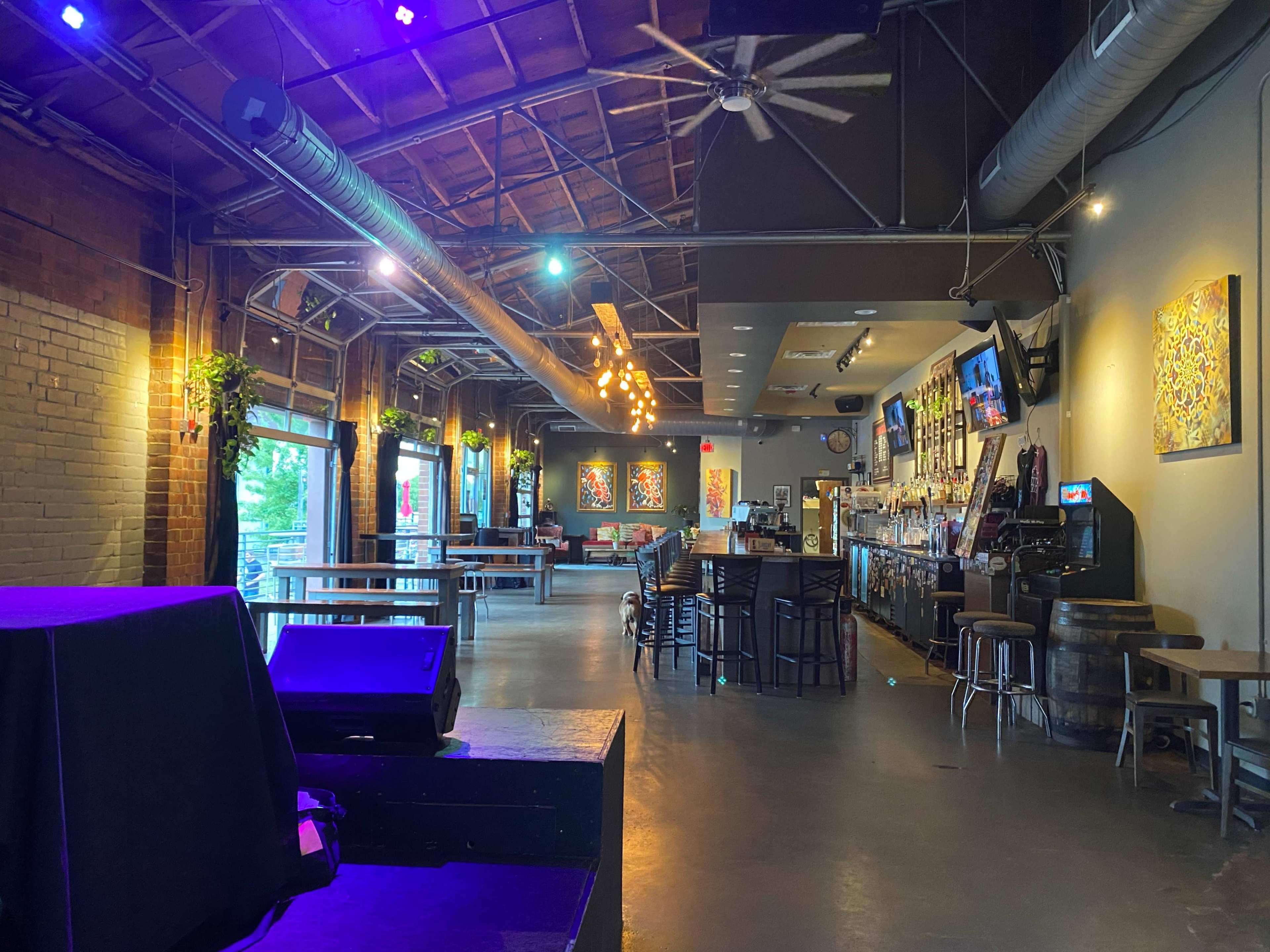 The interior of a spacious bar with a high ceiling, featuring a main seating area with tables, a bar counter, and colorful artwork on the walls.