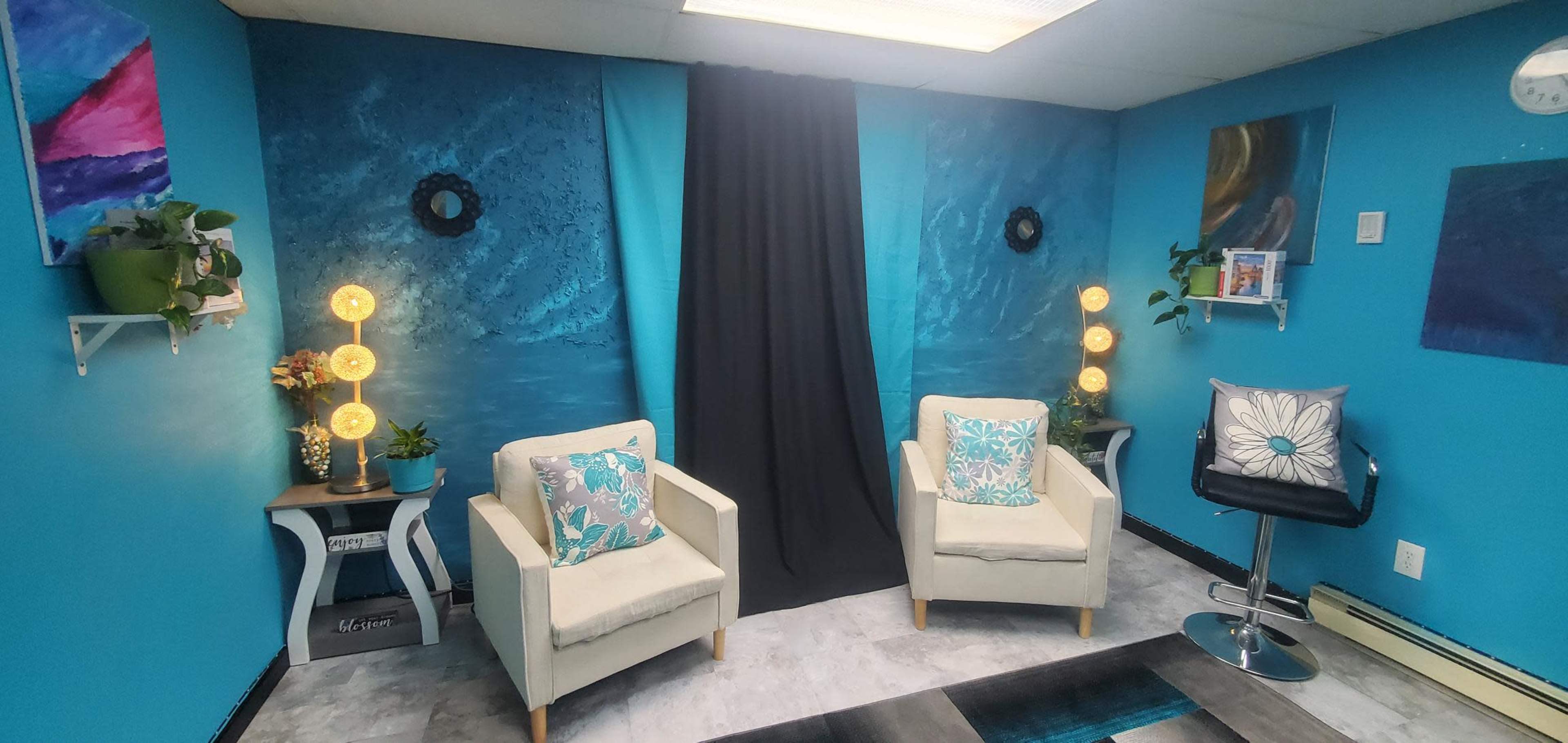 The scene features a brightly painted blue room with two beige armchairs, a black curtain, decorative wall art, and plants, illuminated by two lamp stands.
