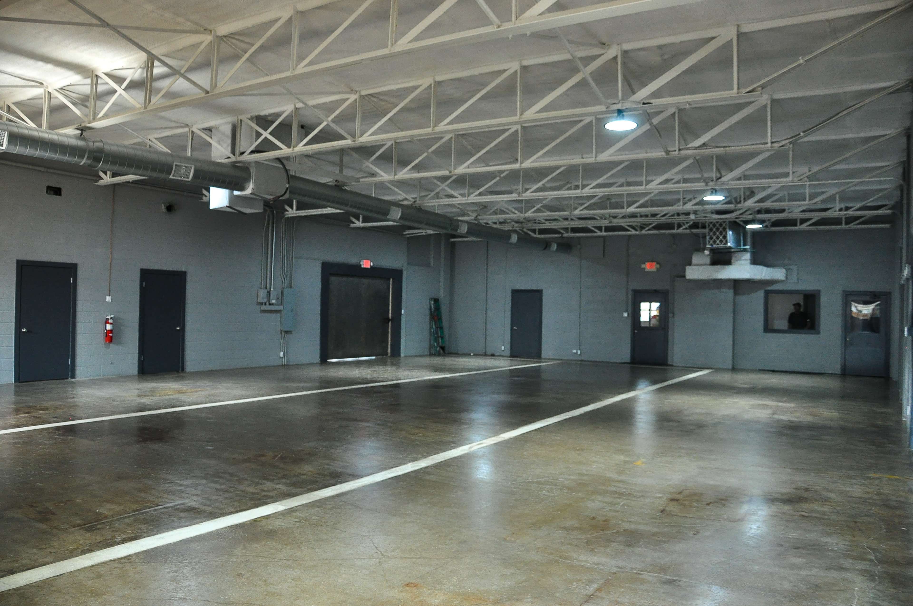 A large, empty industrial space with gray walls, concrete flooring, and several closed doors along the perimeter.