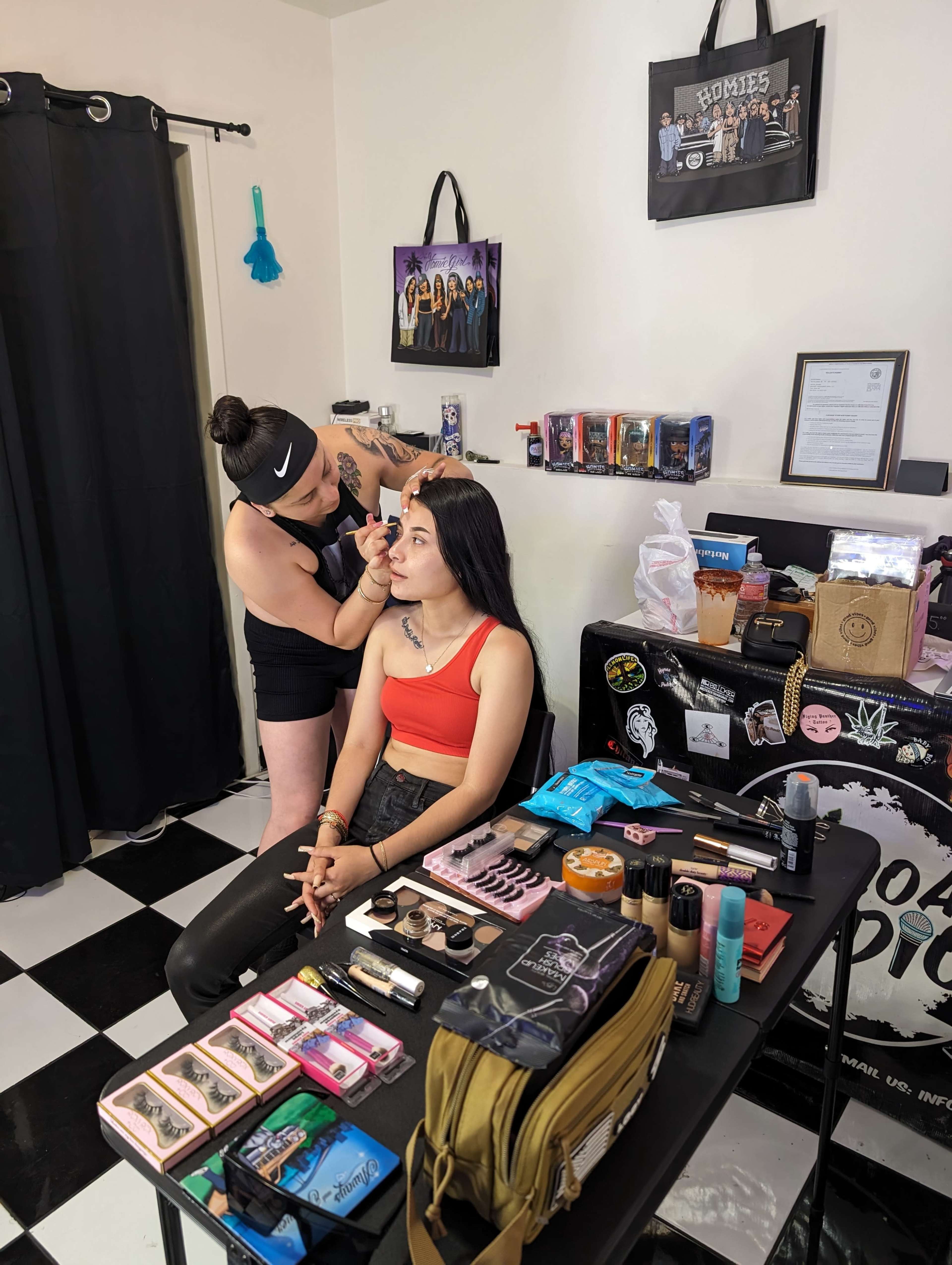 A makeup artist applies makeup to a client at a table filled with various cosmetics in a room with black and white checkered flooring.