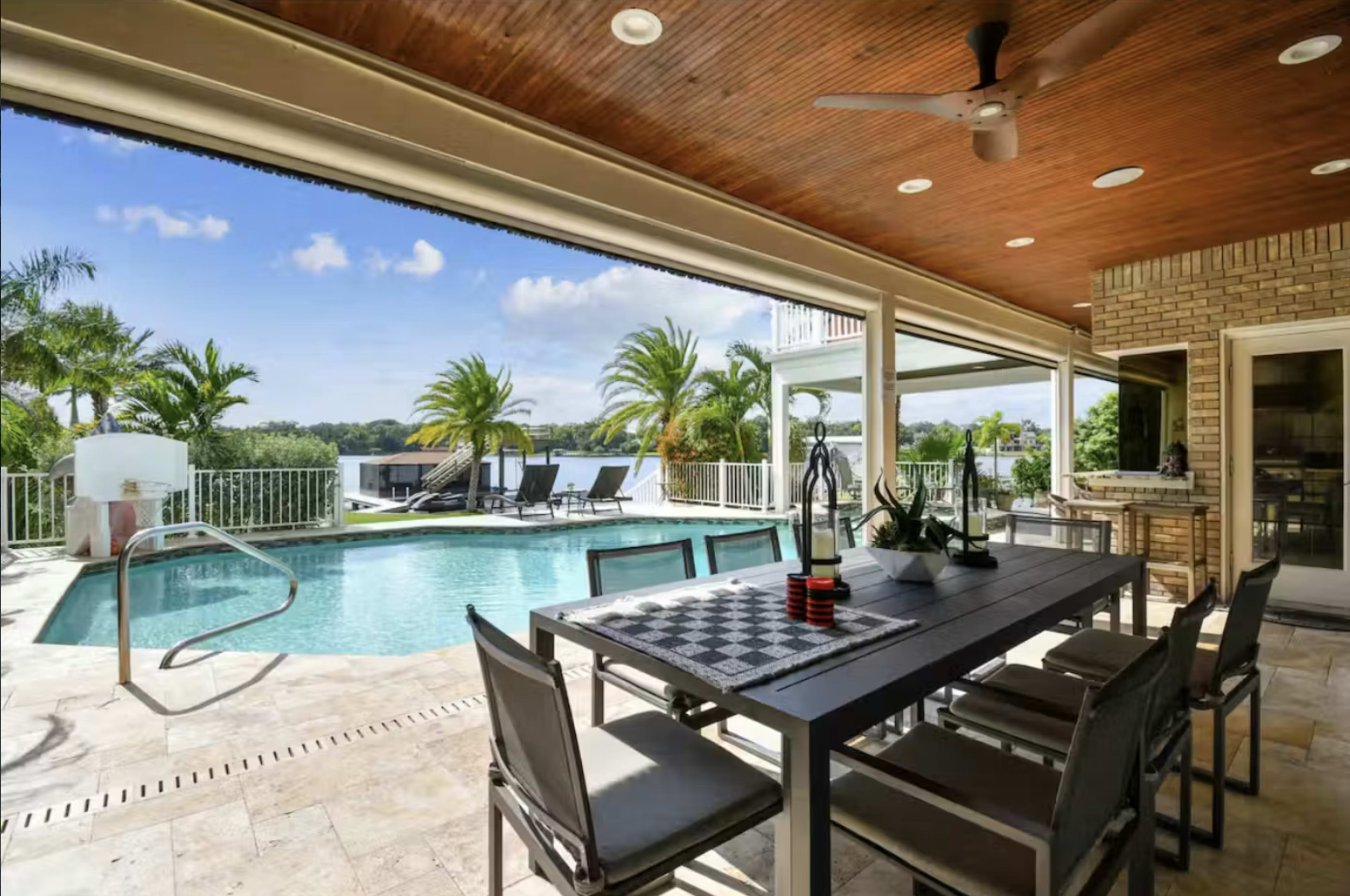 The image shows a patio area with a dining table and chairs overlooking a swimming pool surrounded by palm trees and lounge chairs.