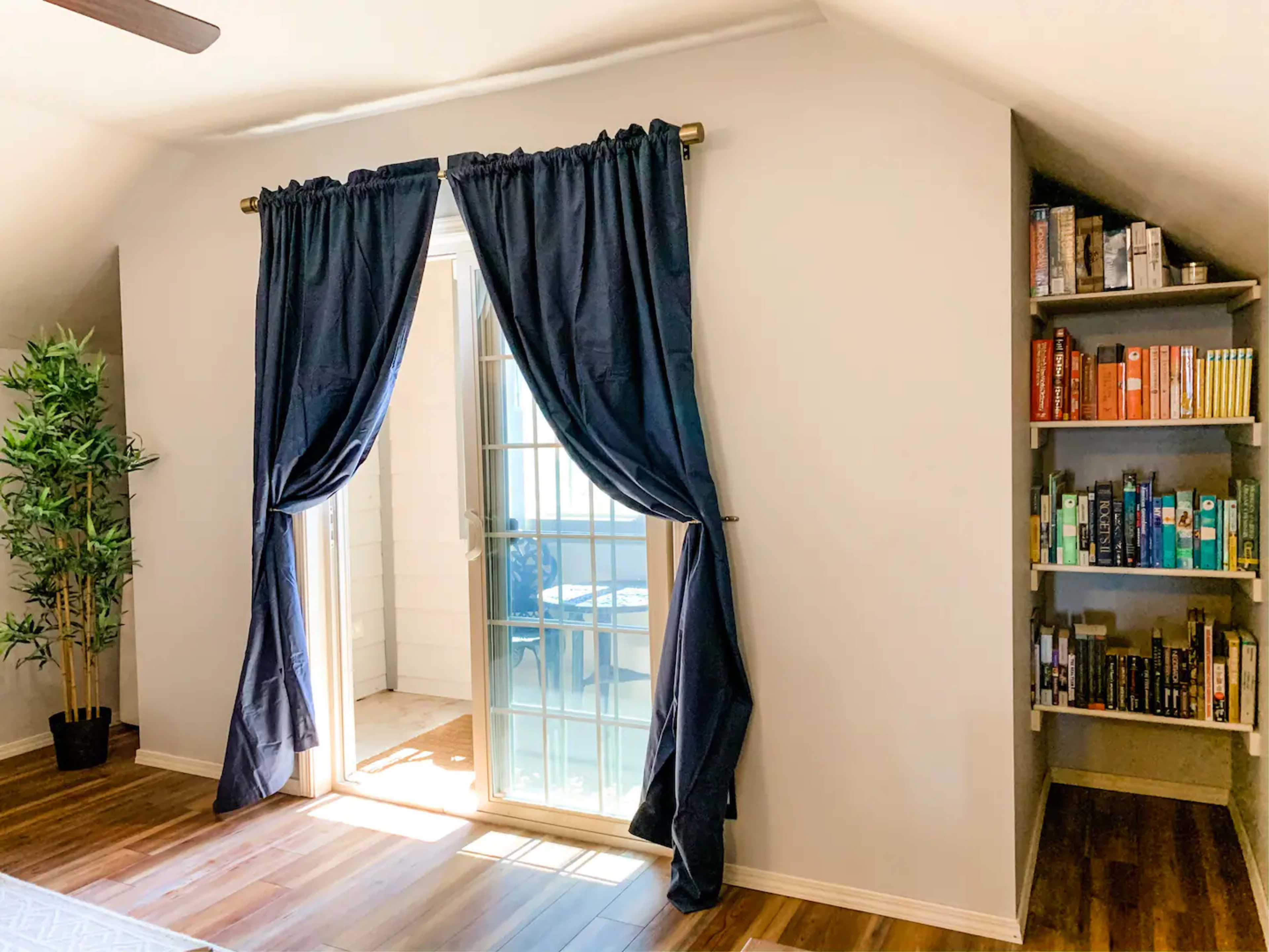 A sunlit room features a sliding glass door with navy curtains and a bookshelf filled with books against a light-colored wall.