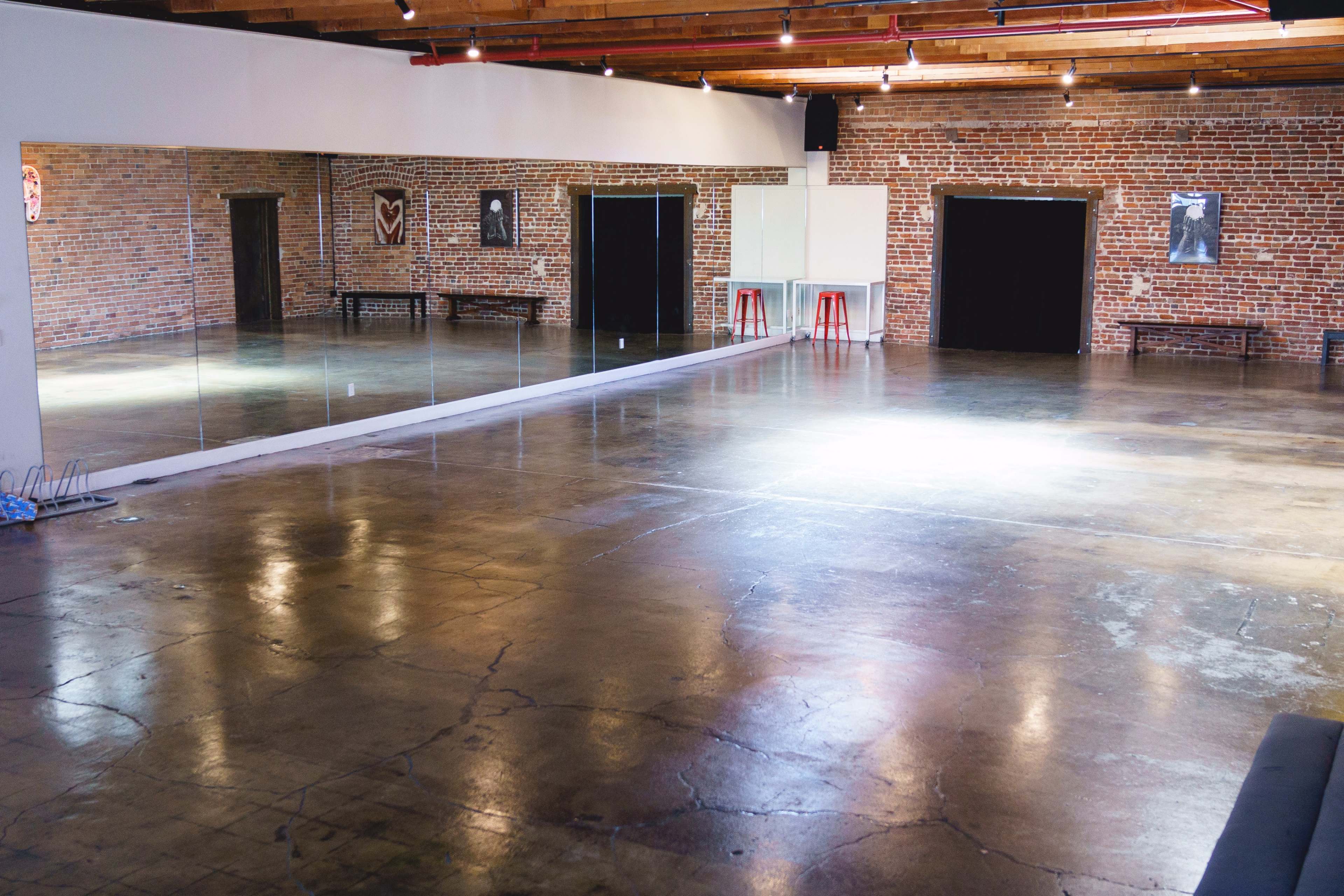 A spacious, empty room with exposed brick walls, polished concrete flooring, and large mirrors along one side.