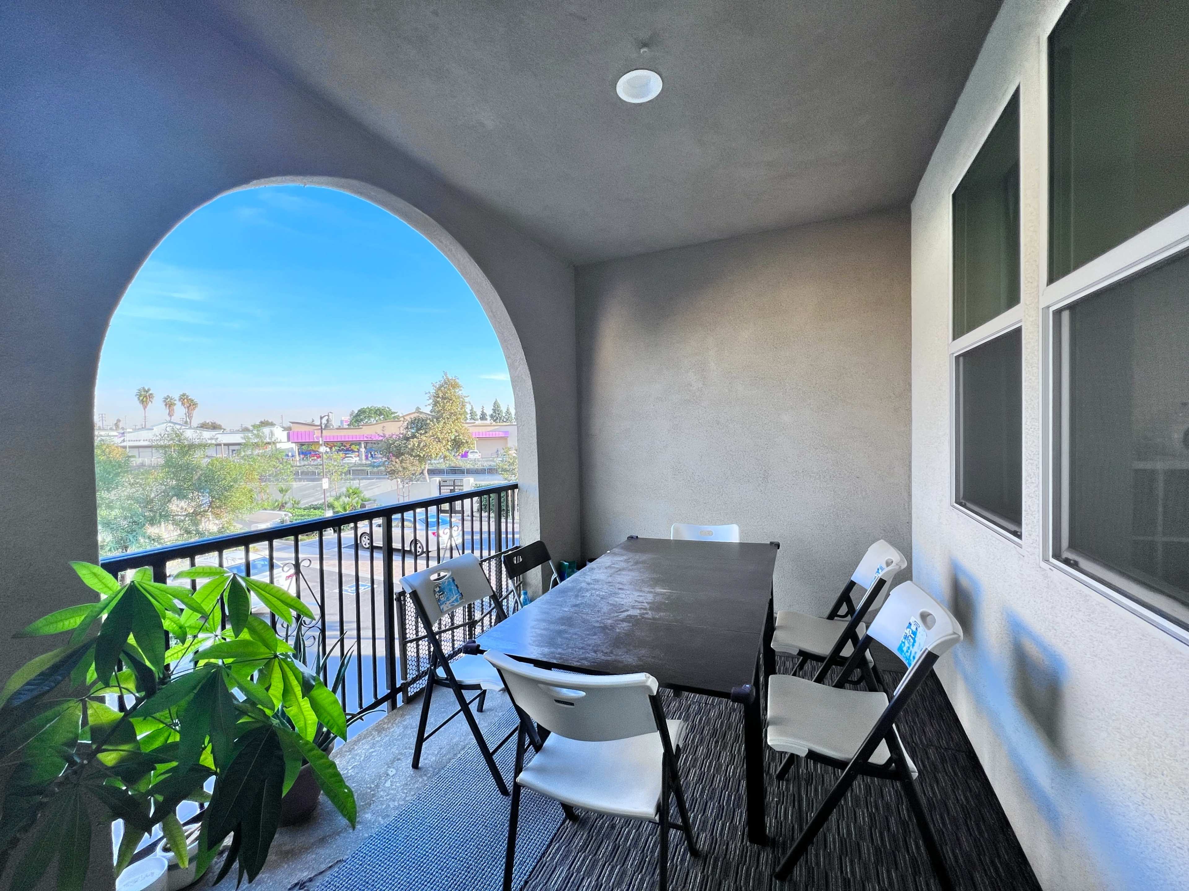 A covered balcony features a rectangular table surrounded by six folding chairs, with a view of palm trees and buildings outside.