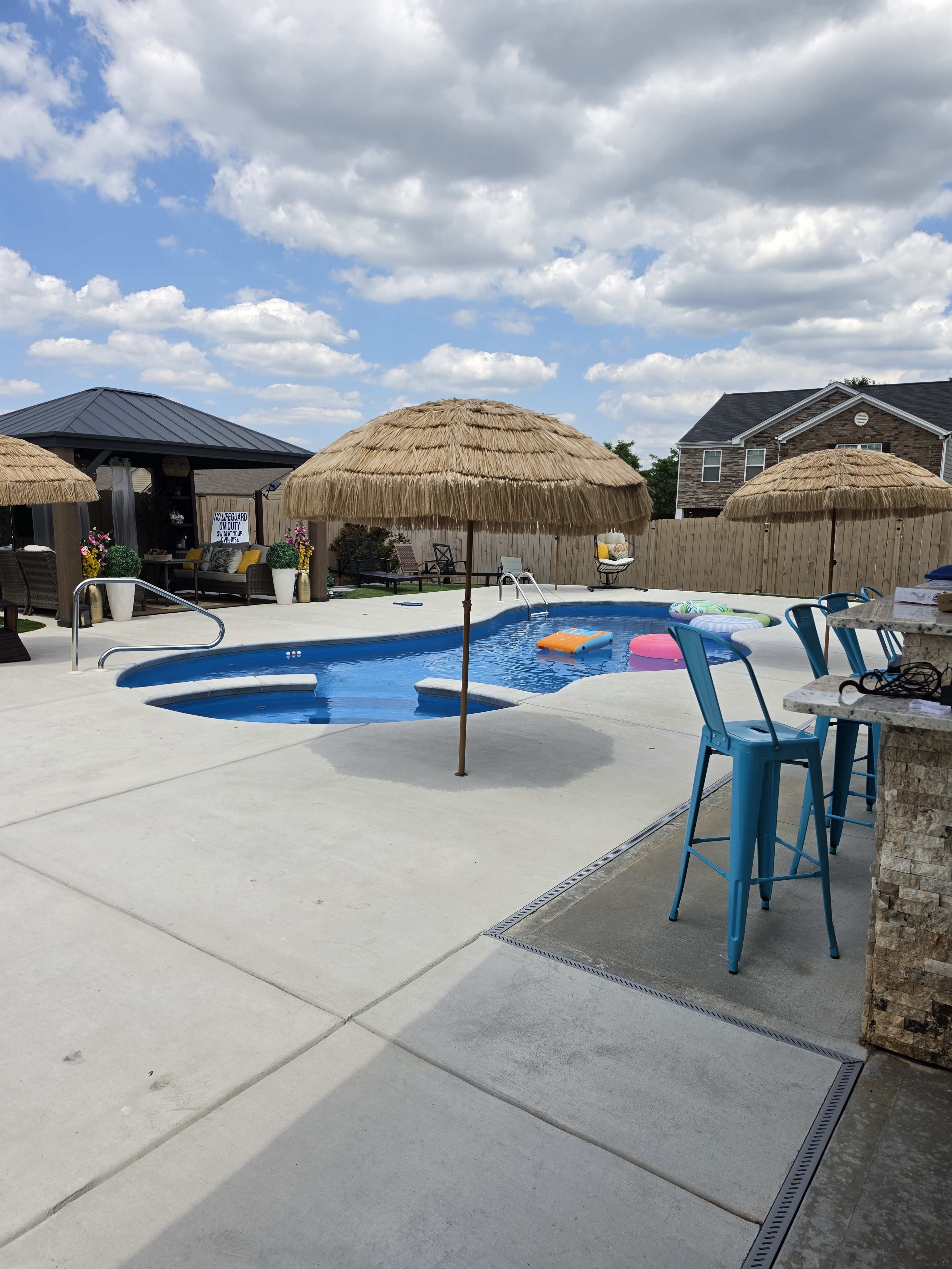 The image shows a swimming pool with thatched umbrellas, colorful floaties, and seating areas in a backyard setting.