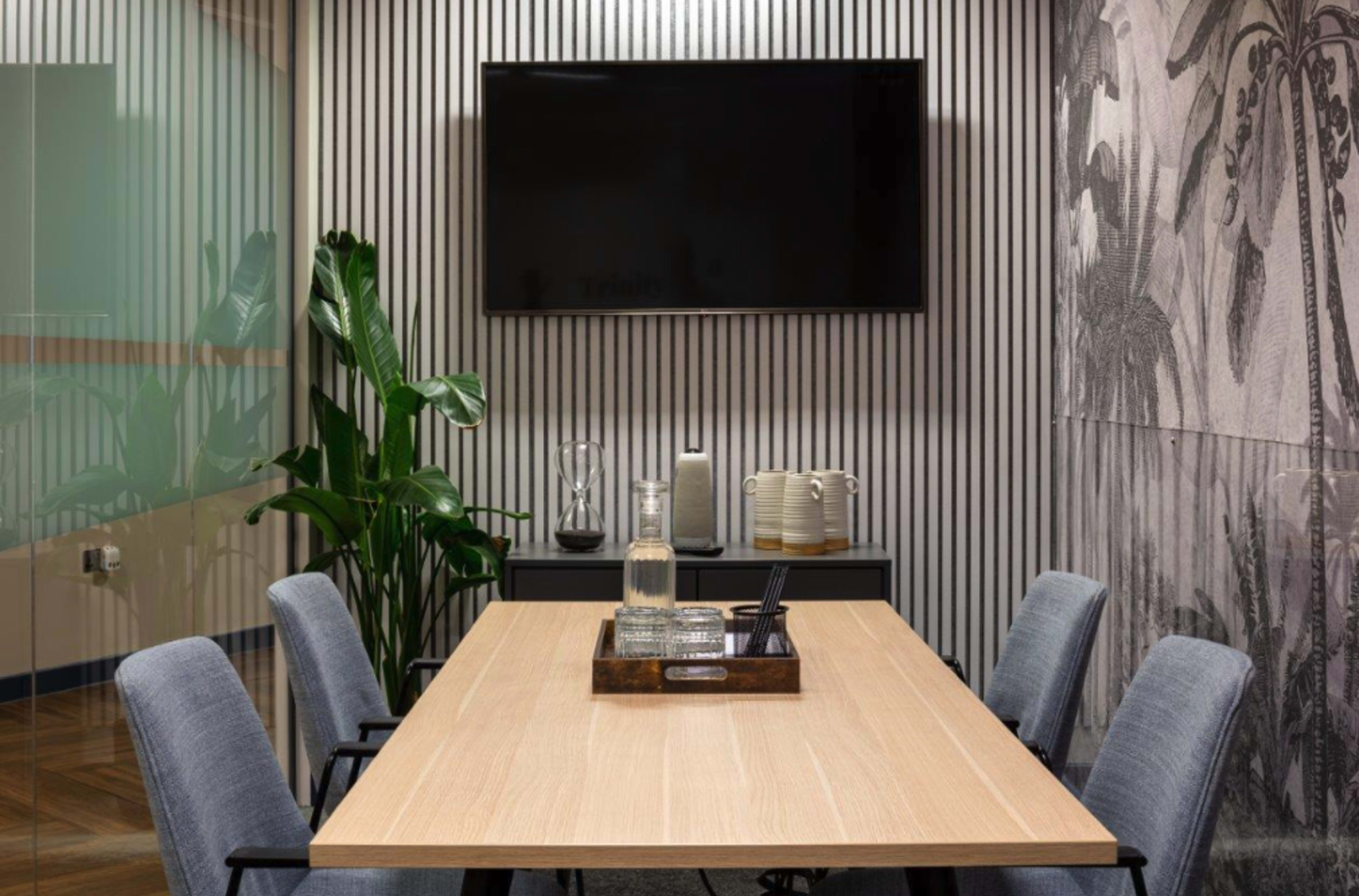 A modern meeting room features a wooden table surrounded by gray chairs, a wall-mounted television, and decorative plants.
