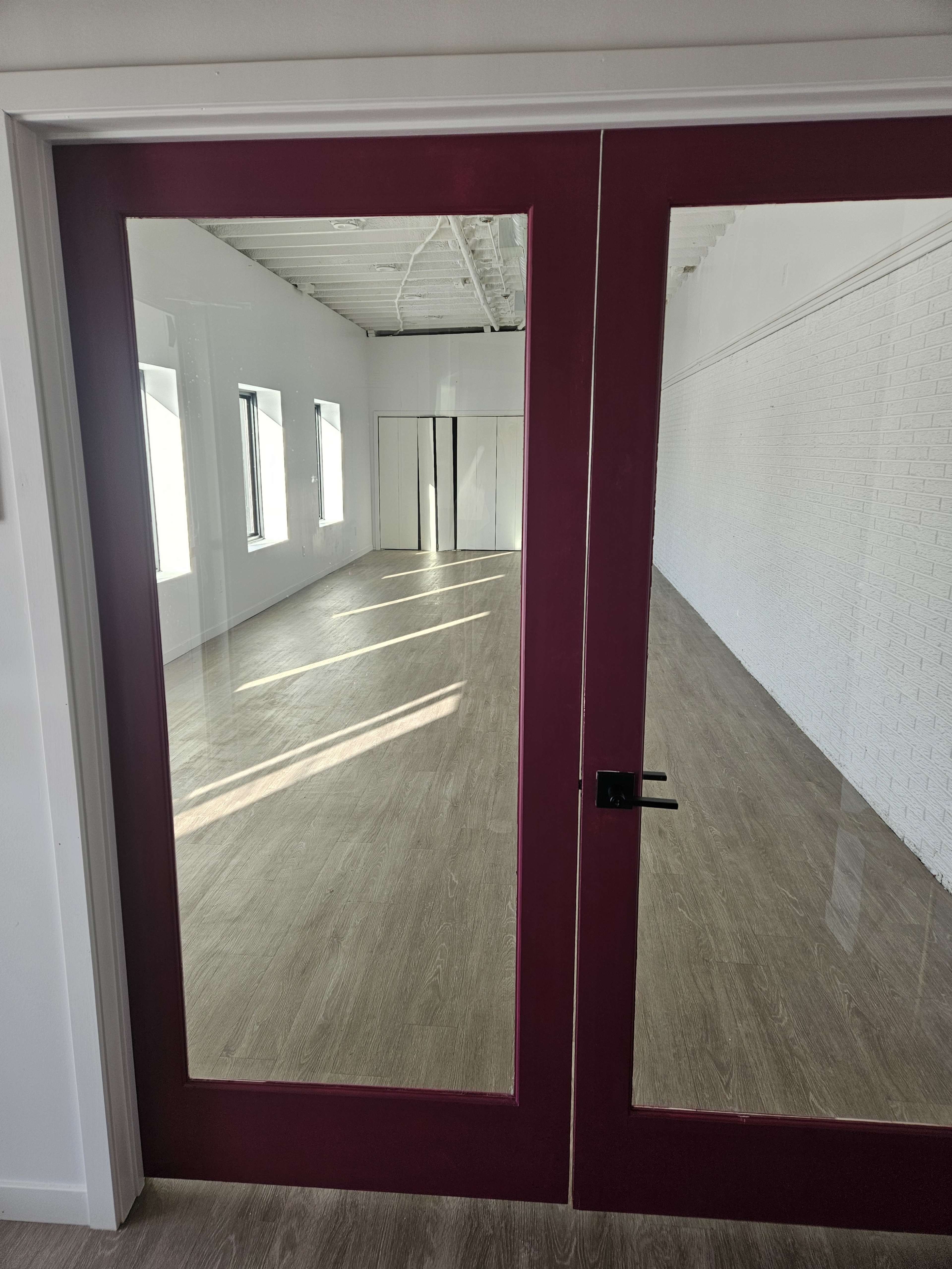 The image shows a pair of glass doors with maroon frames opening into a bright, empty room with wooden flooring and white walls.