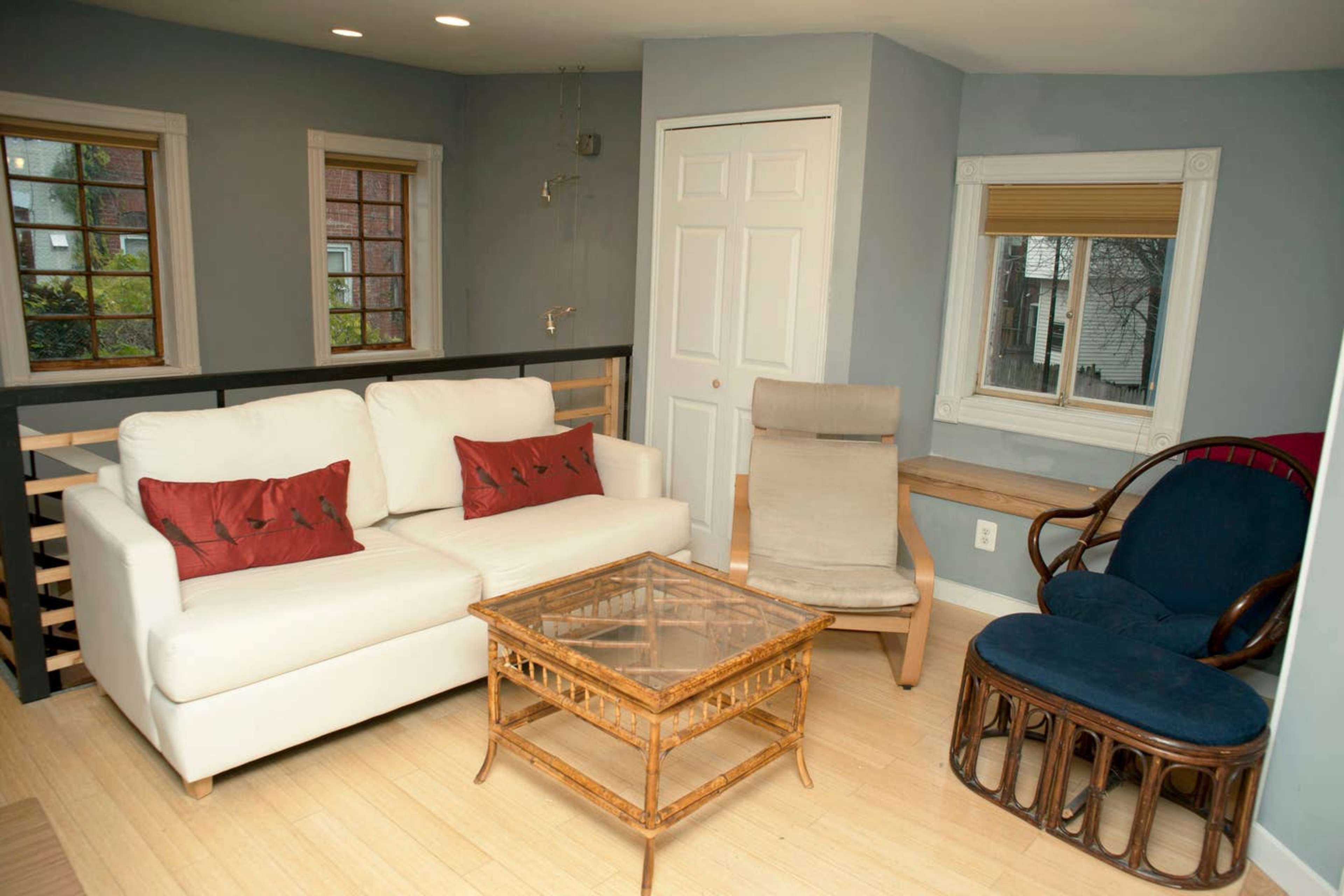 The image shows a living room with a white couch, a glass coffee table, a wooden chair, and two windows allowing natural light.