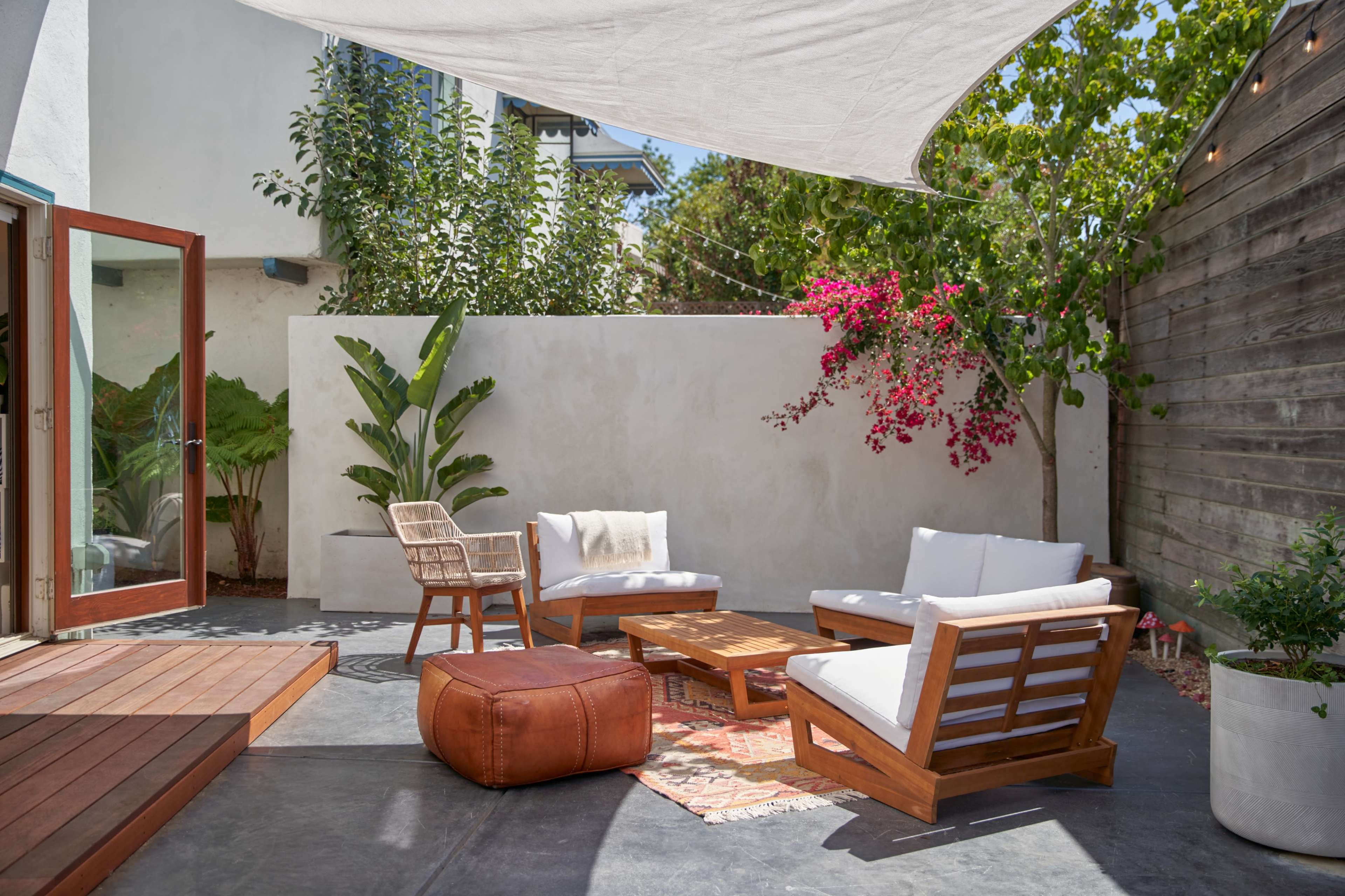 A small outdoor lounge area features wooden furniture, a coffee table, and a large shaded canopy, surrounded by plants and a white wall with flowering vines.