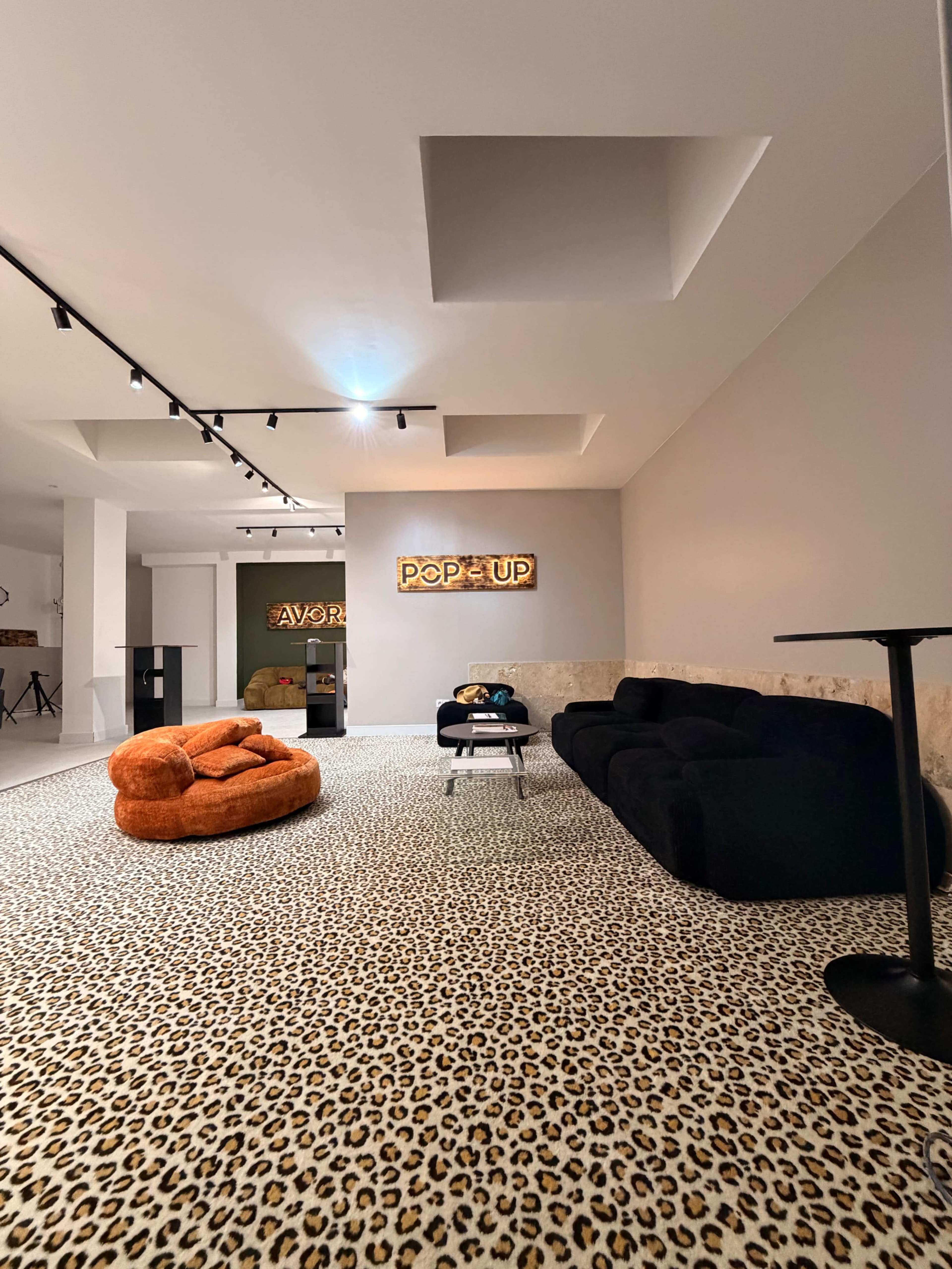 The image shows a modern interior space featuring a leopard-print carpet, black sofas, an orange bean bag chair, and neon signs on the walls.