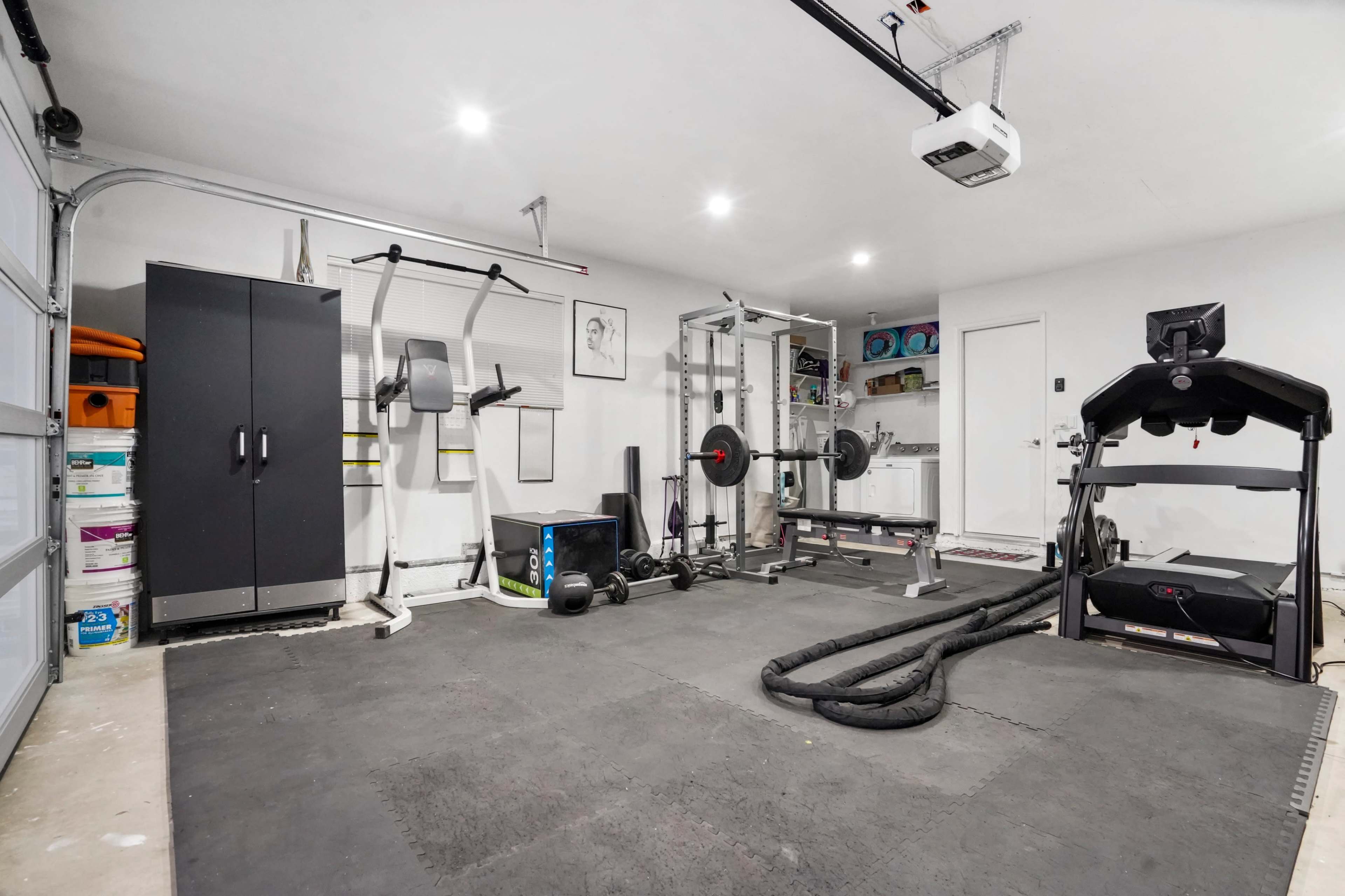 A home gym with exercise equipment including a treadmill, weightlifting bench, weights, and mats arranged in a garage space.