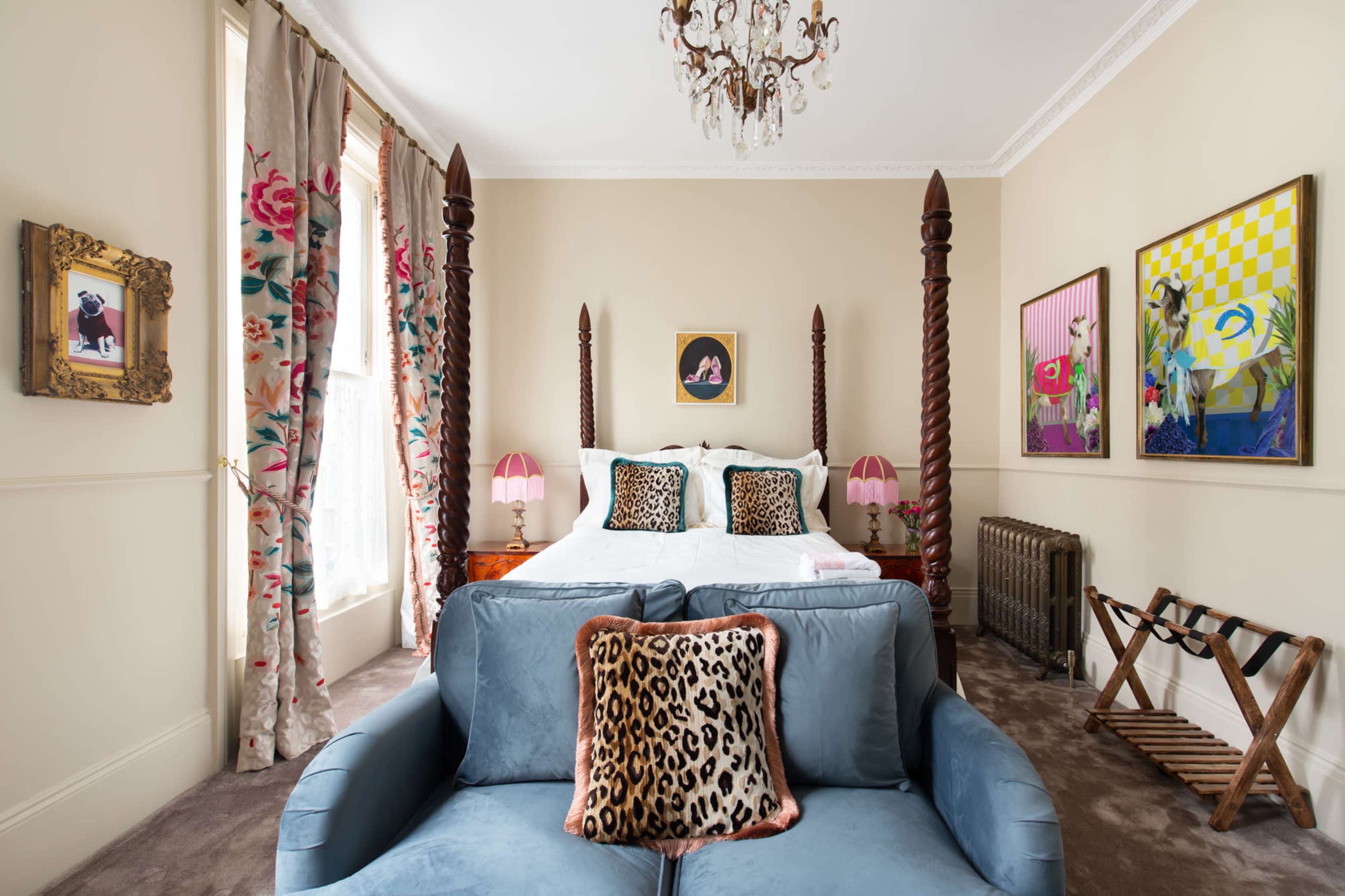 A bedroom features a king-size bed with tall wooden posts, a couch with leopard-print pillows, and colorful artwork on the walls.
