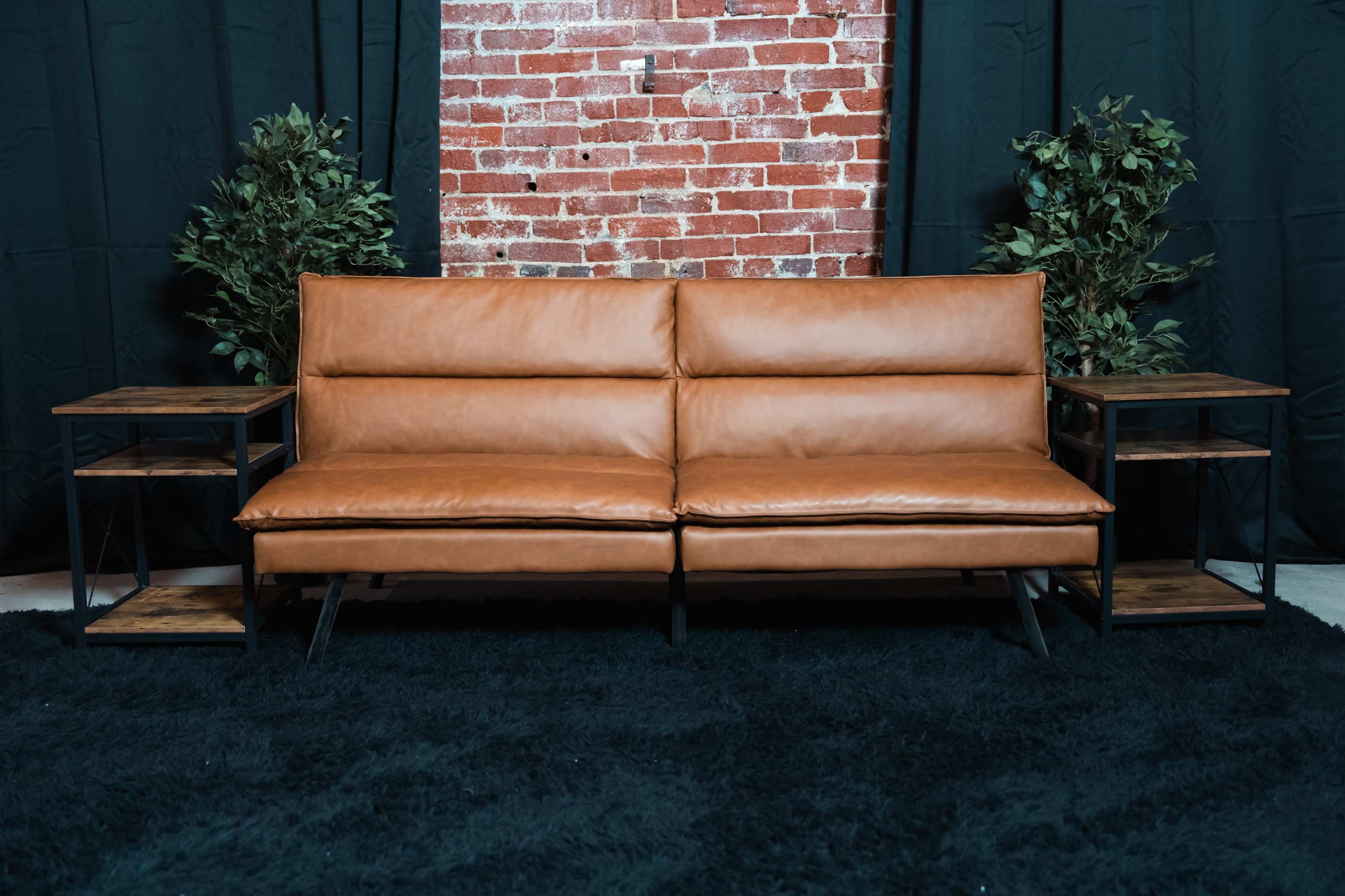 A brown leather sofa is positioned against a brick wall, flanked by two wooden side tables on either side and surrounded by dark curtains and potted plants.