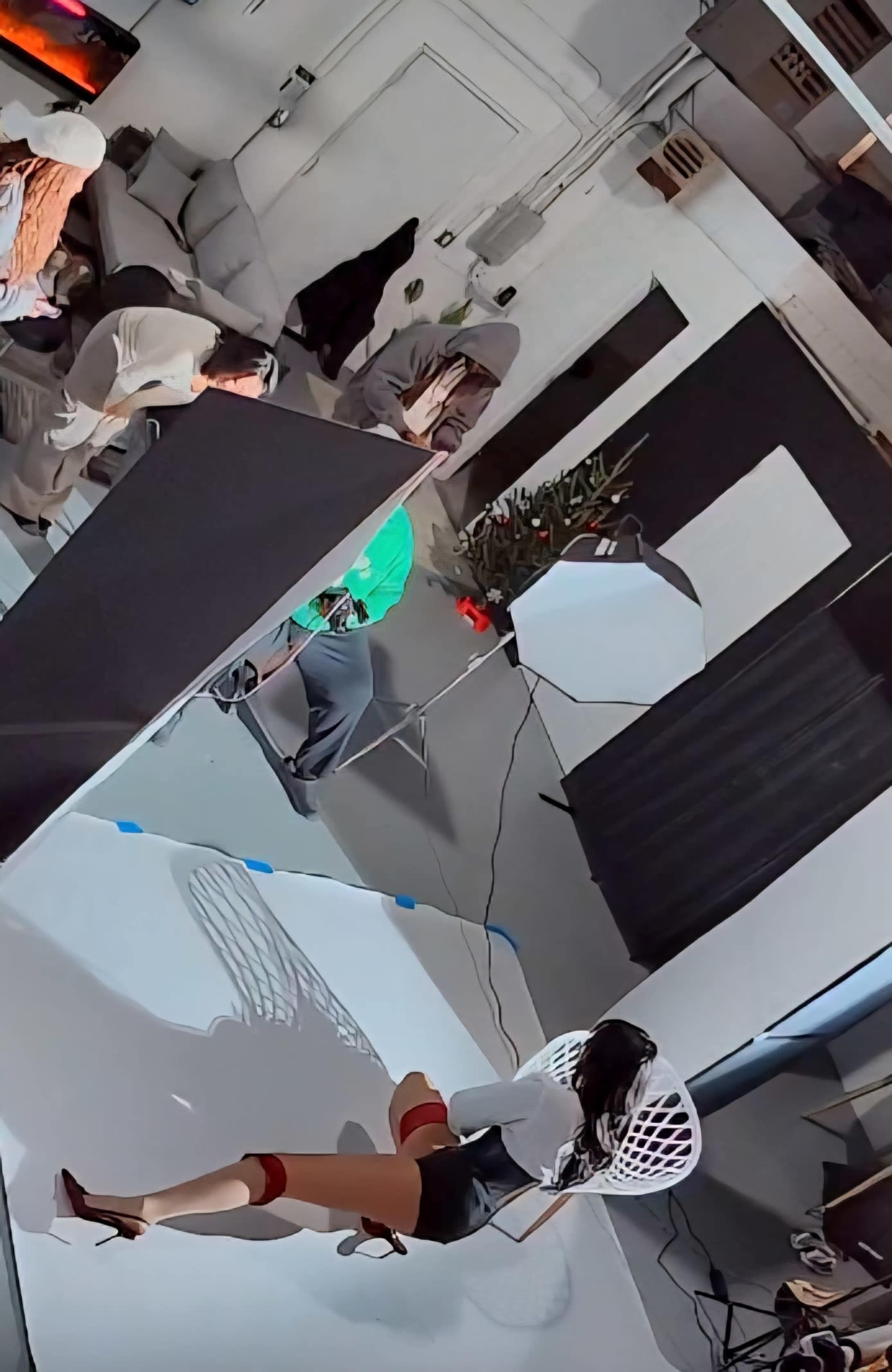 A photoshoot is taking place in an indoor studio, featuring a model in a chair and several people working in the background.
