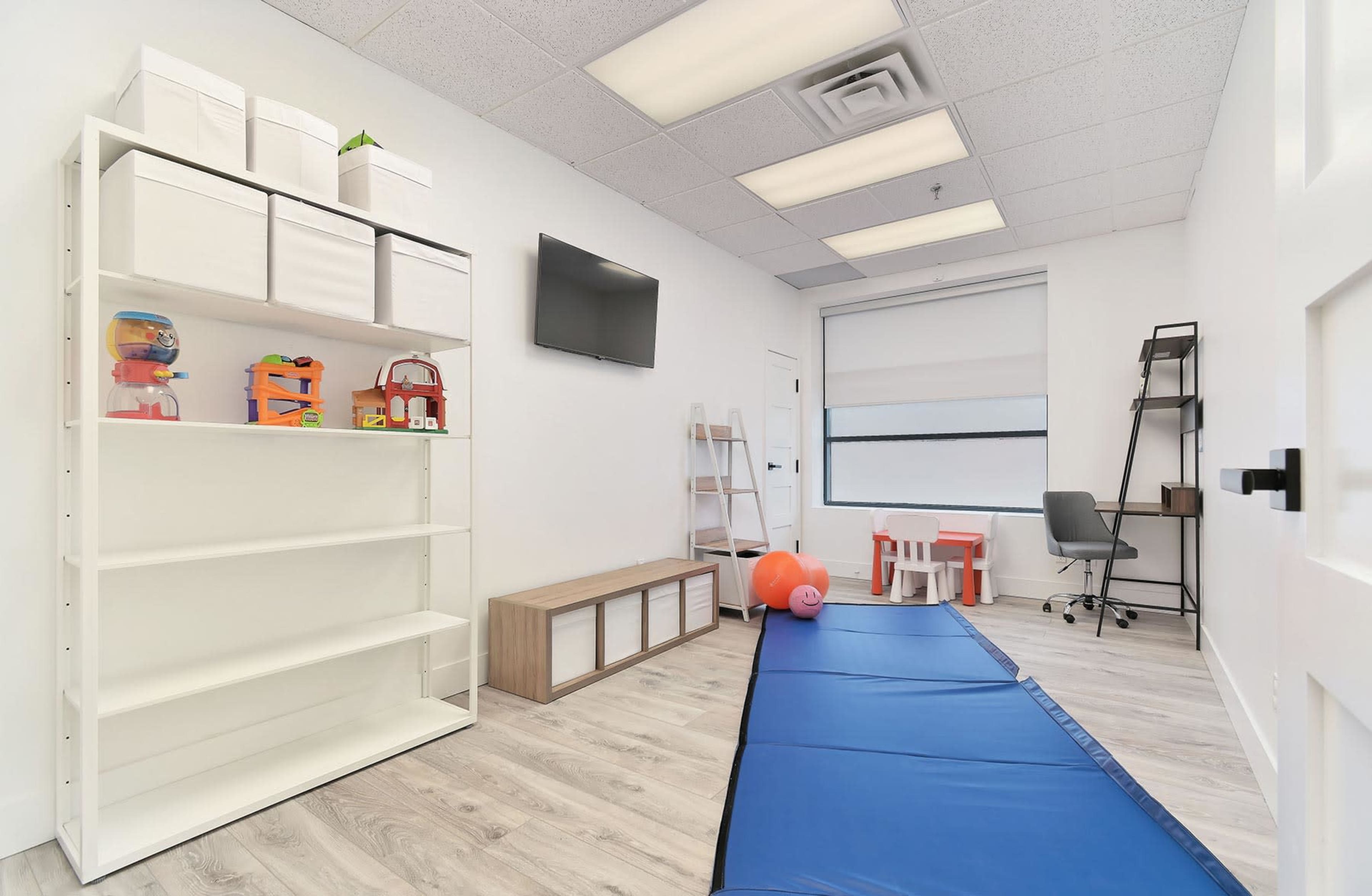 A bright, organized room featuring a shelf with toys, a TV on the wall, a desk with a chair, and a padded blue mat on the floor.