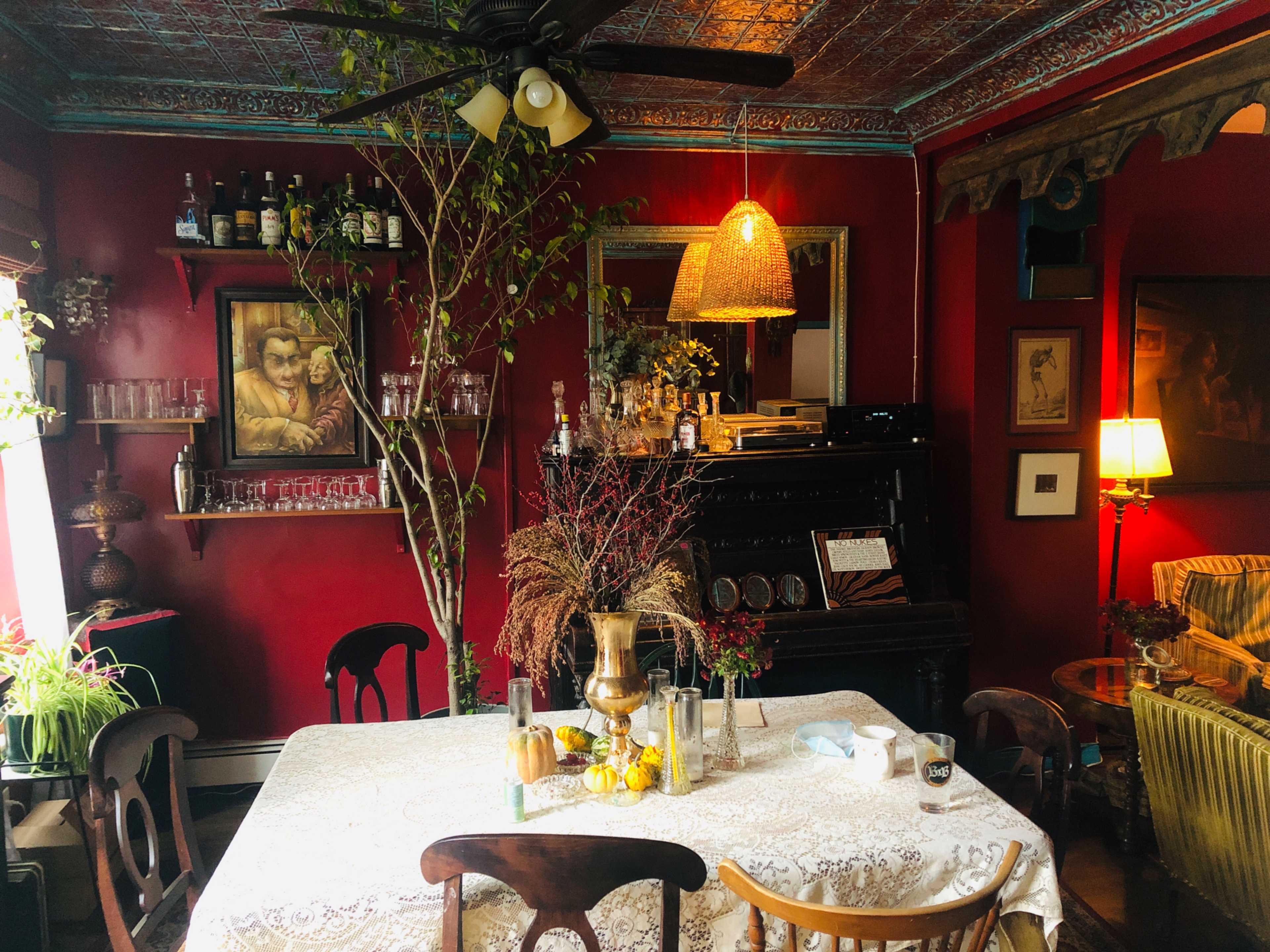 The image shows a cozy dining area with a round table set with a lace tablecloth, surrounded by wooden chairs, and featuring a piano in the background along with various plants and vintage decor.