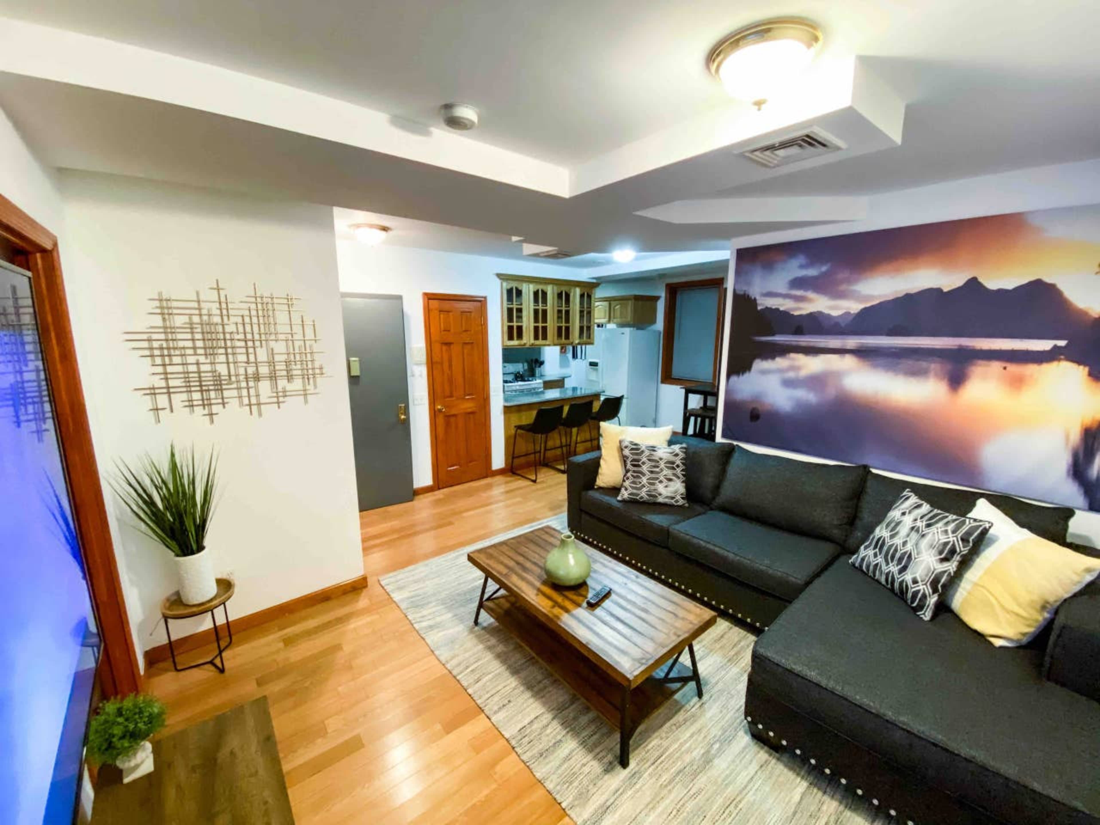 A modern living room features a black sectional sofa, wooden coffee table, and a large wall mural of a mountain landscape, with a dining area visible in the background.