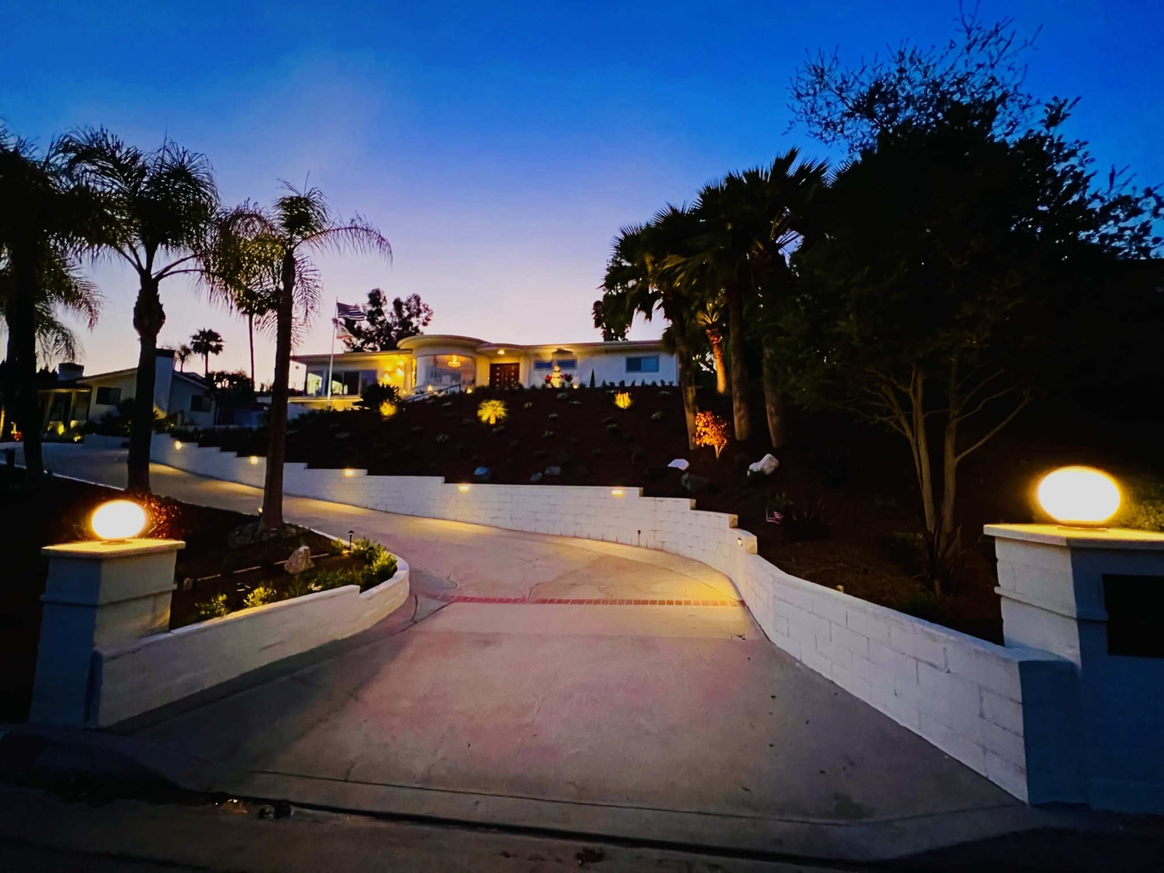 A well-lit driveway leads up to a modern house surrounded by palm trees and landscaping against a twilight sky.