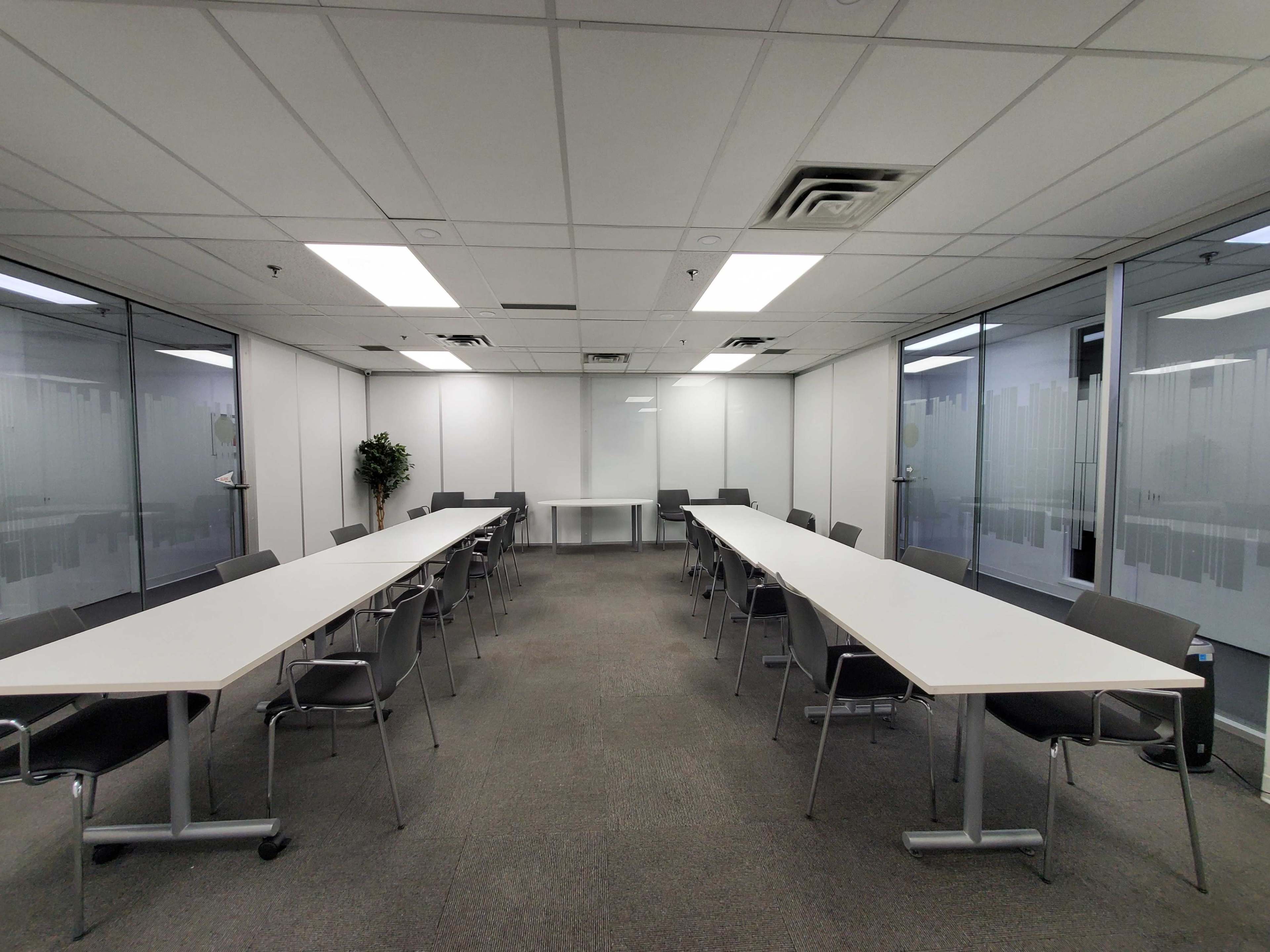 A spacious conference room features two long tables arranged in a U-shape, surrounded by chairs and fitted with glass walls and a projector screen at one end.