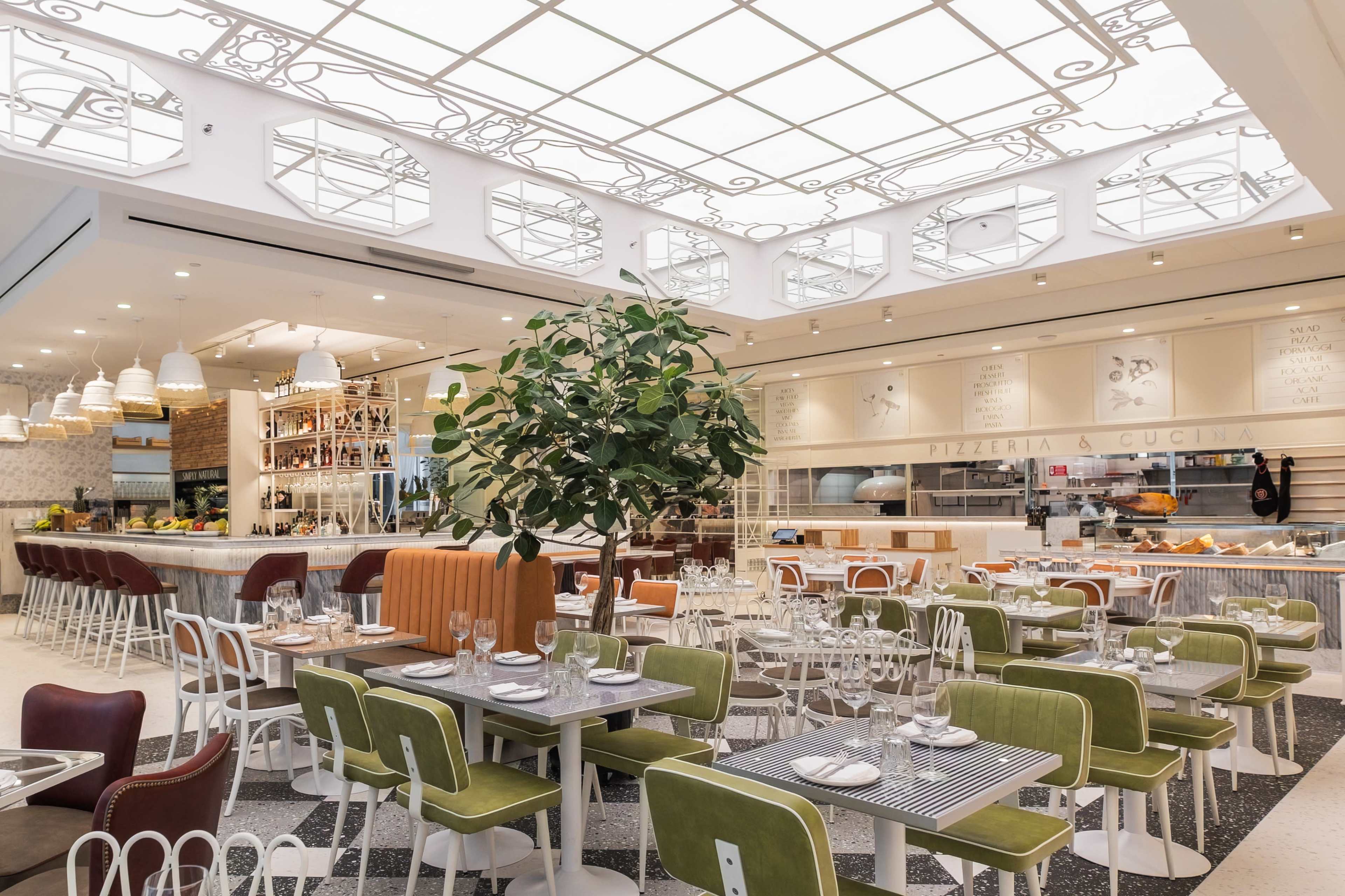 The image shows a modern restaurant interior with a large glass roof, dining tables, green chairs, and a central tree.