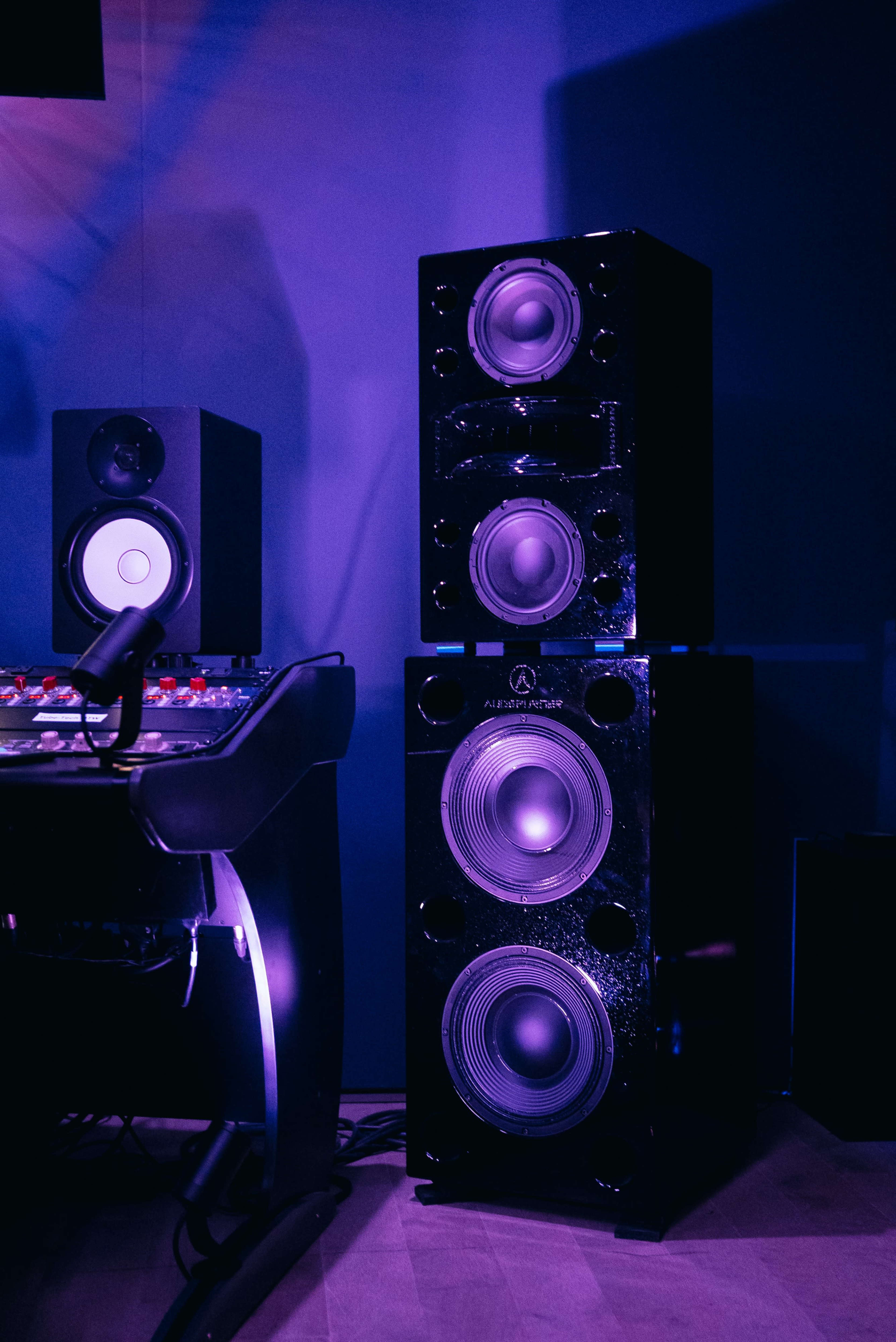 The image shows a tall stack of black audio speakers beside a music production console in a dimly lit studio.